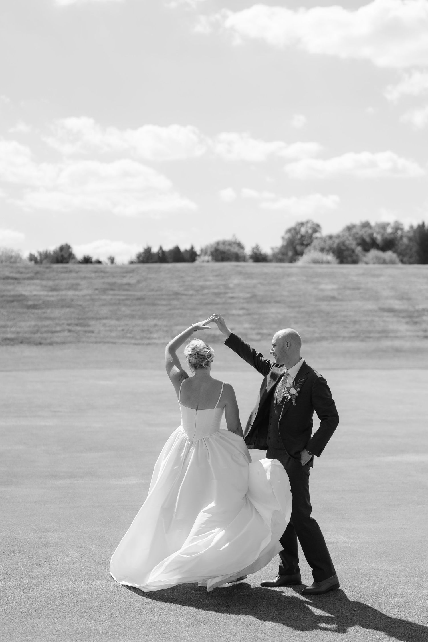 Black and white photo of groom spinning bride in flowing wedding gown on open golf course under cloudy sky.