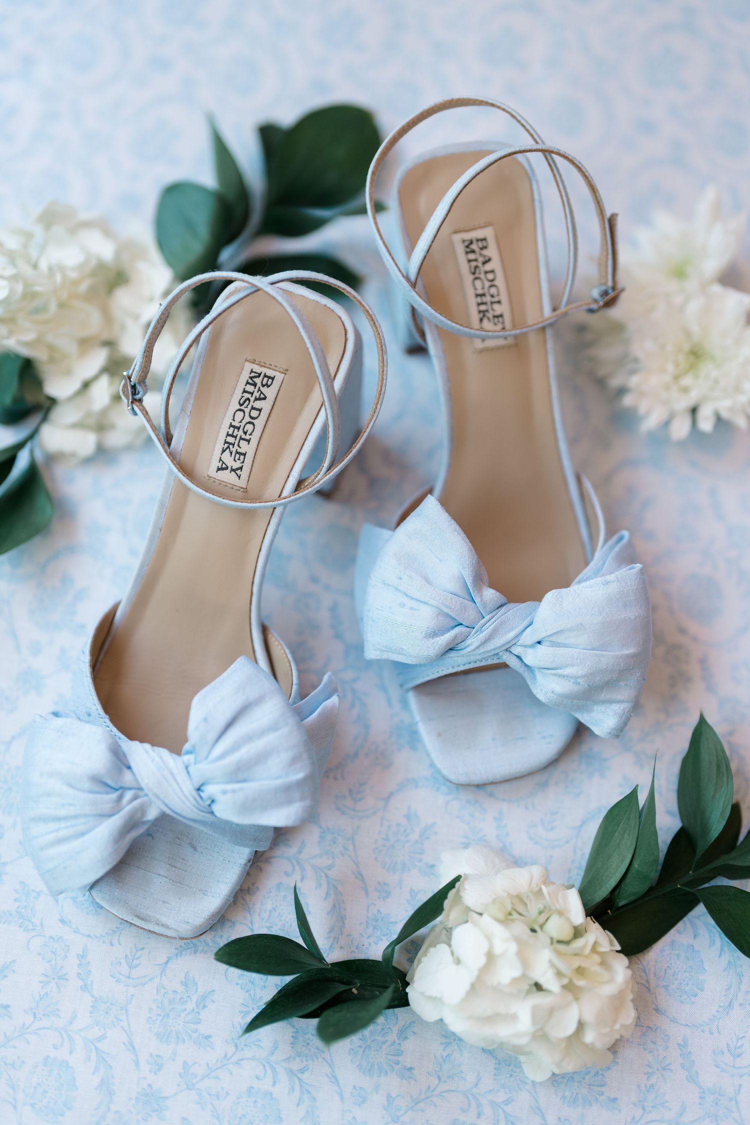 Light blue Badgley Mischka bridal heels with bow detail surrounded by white flowers and greenery.