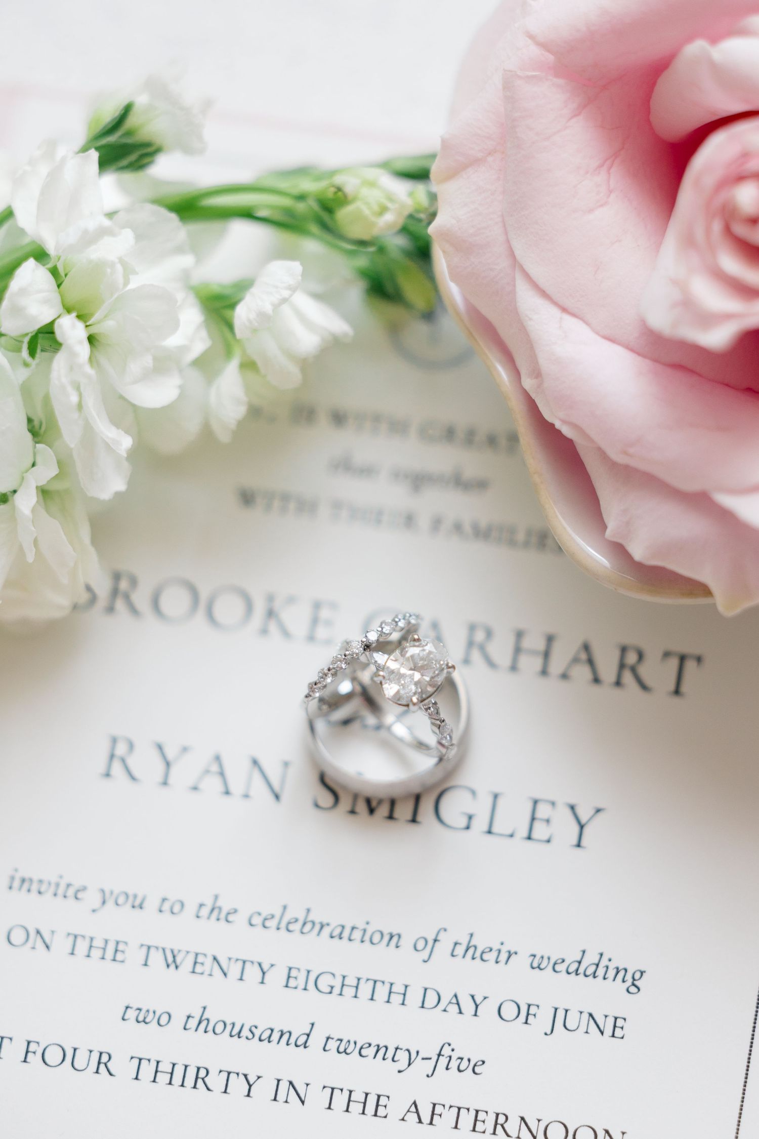 Elegant wedding invitation with pink rose, white flowers and diamond engagement ring on display.