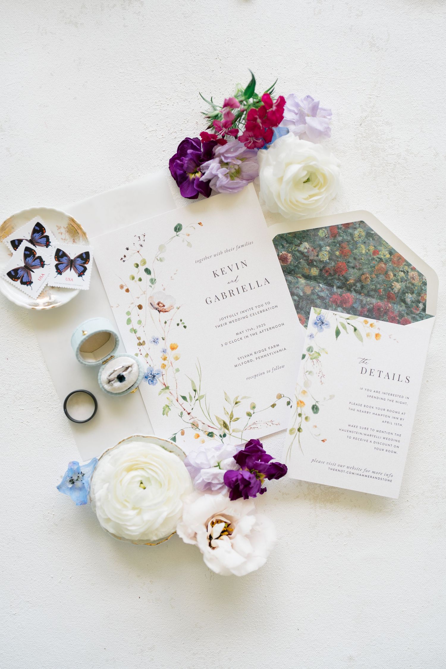 Floral wedding invitation suite with white ranunculus flowers, butterfly decor, and rings styled on light background.