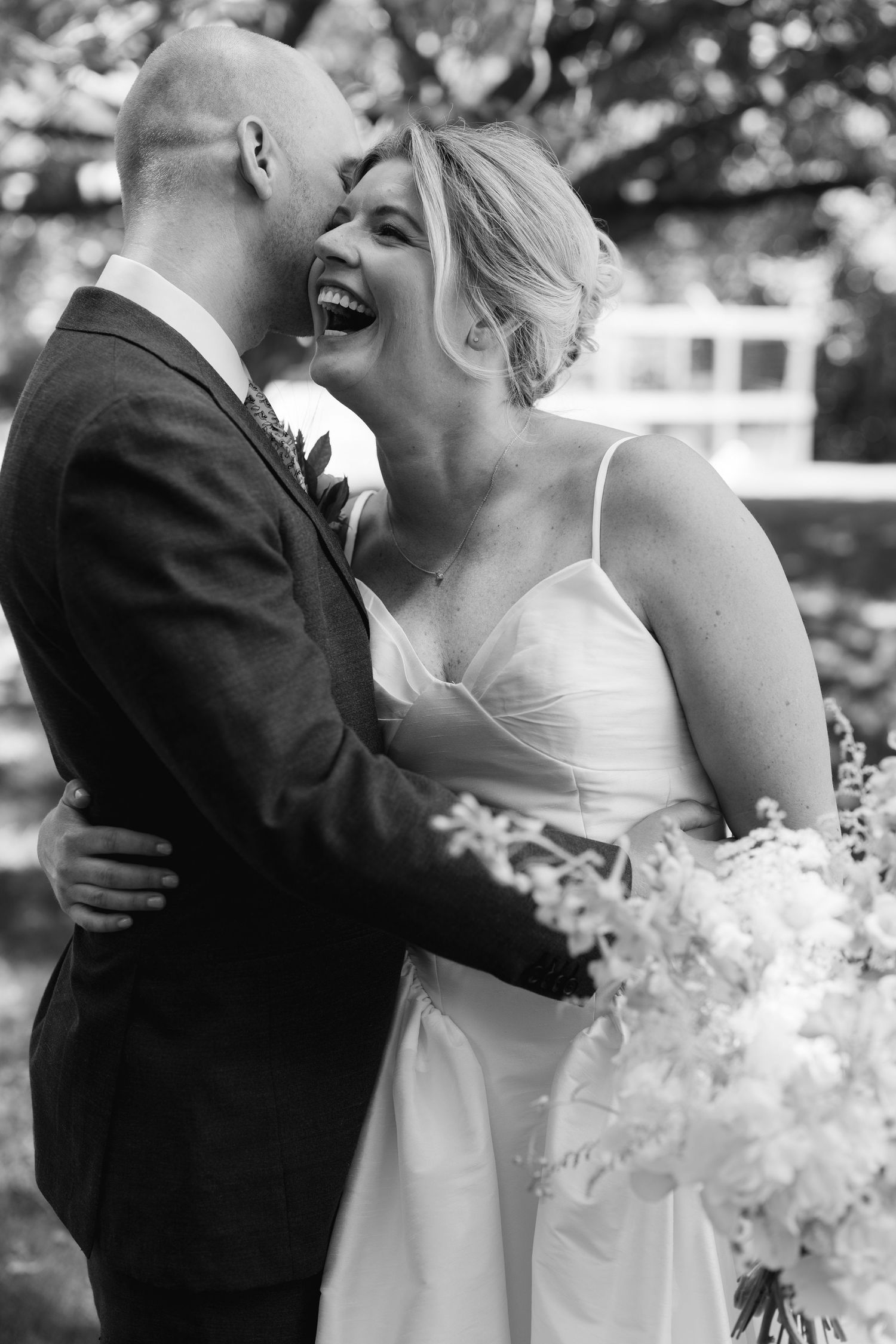 Black and white photo of a laughing bride and groom embracing outdoors on their wedding day.