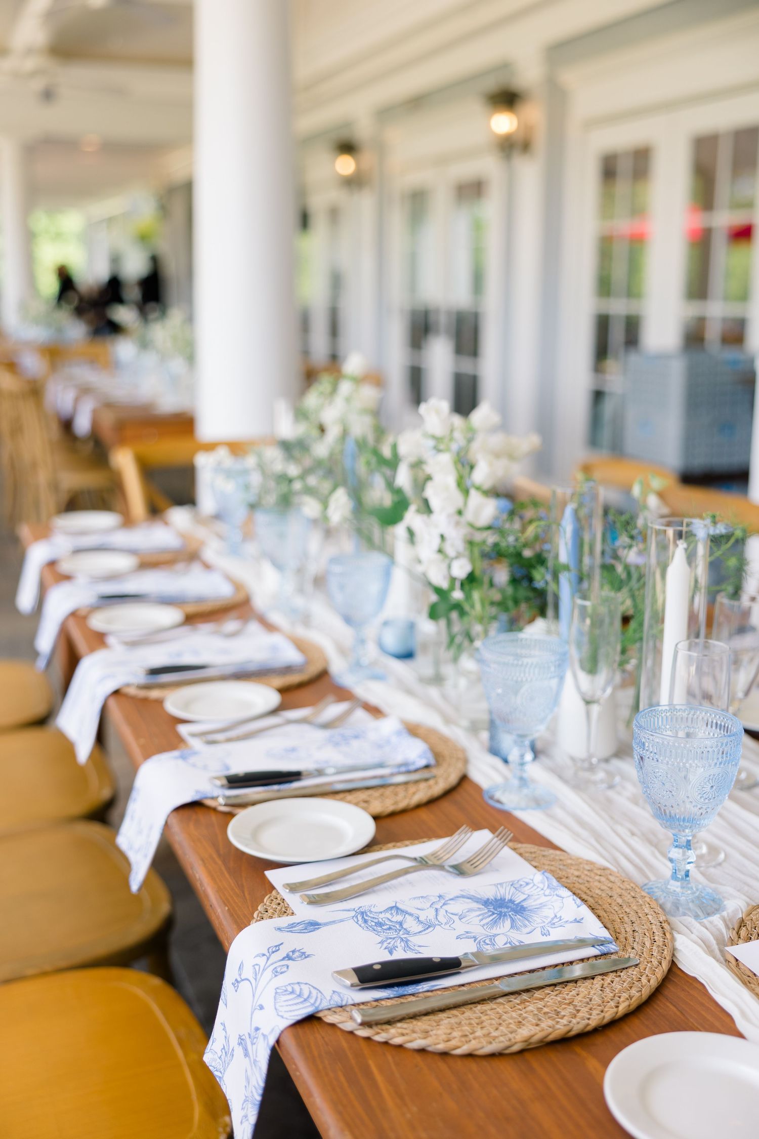 Elegant outdoor wedding reception table with blue glassware, wicker chargers, and white floral centerpieces.