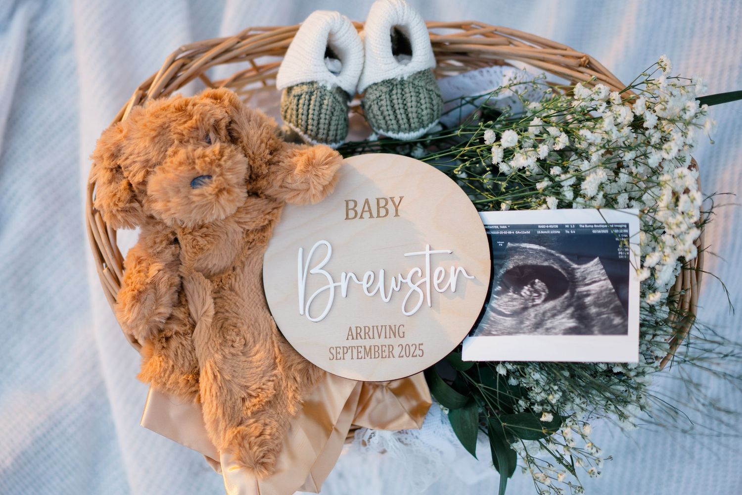 Baby announcement display with ultrasound photo, wooden name disc, teddy bear and baby's breath flowers in wicker basket.