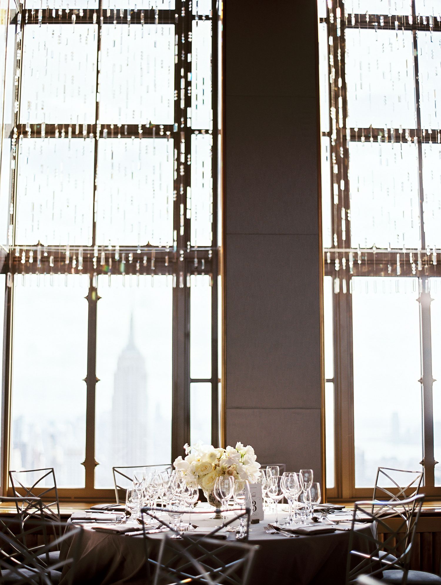 Elegant table setting with white floral centerpiece against tall windows in an upscale dining room overlooking a city view.