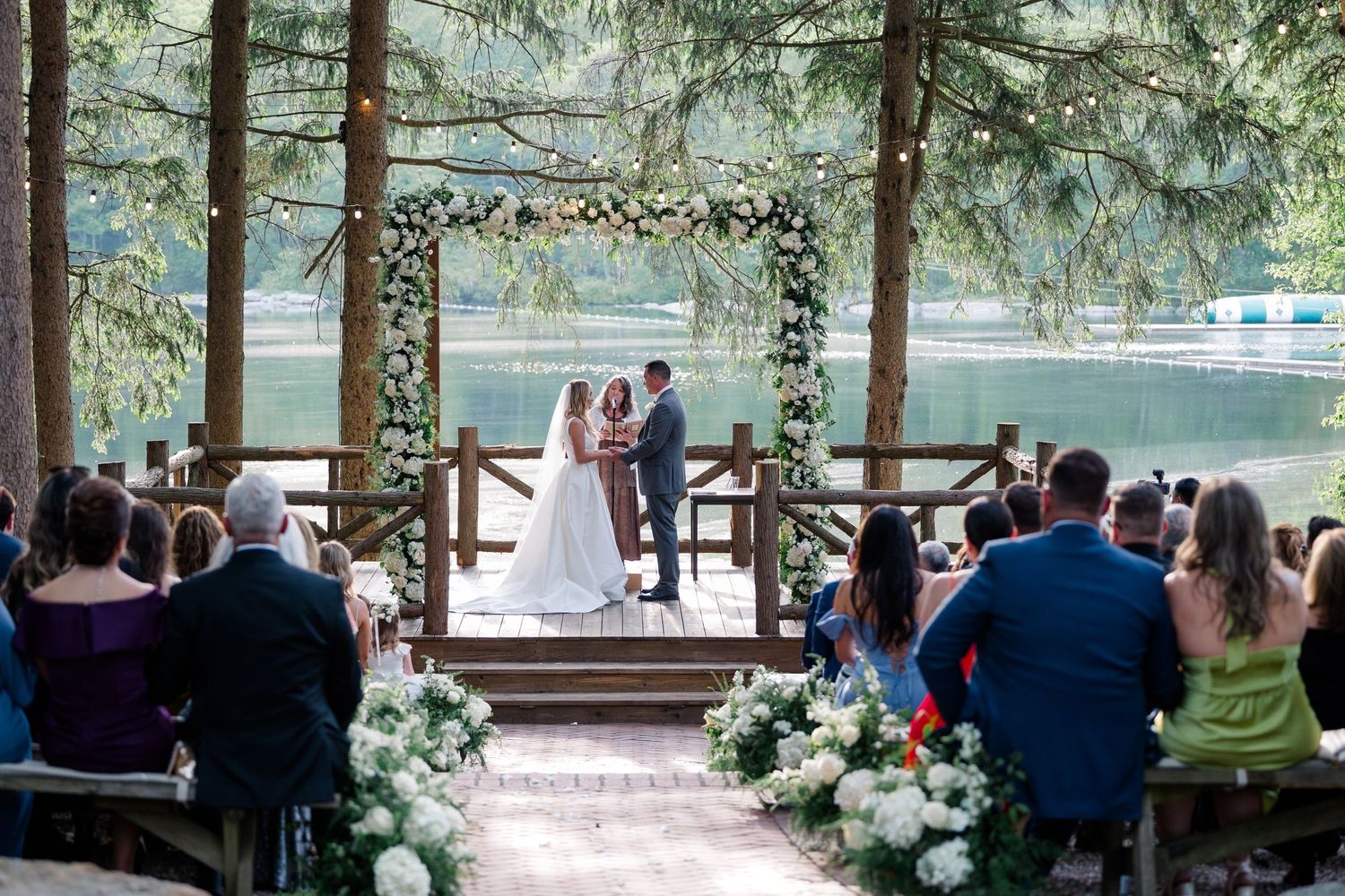 An outdoor wedding ceremony under a floral arch by a lake surrounded by evergreen trees and seated guests.