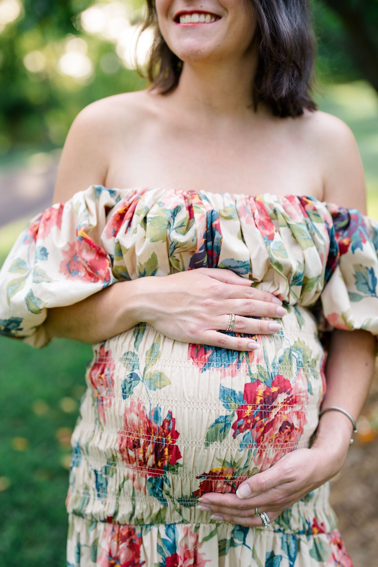 Smiling pregnant woman in floral off-shoulder dress cradling baby bump outdoors.