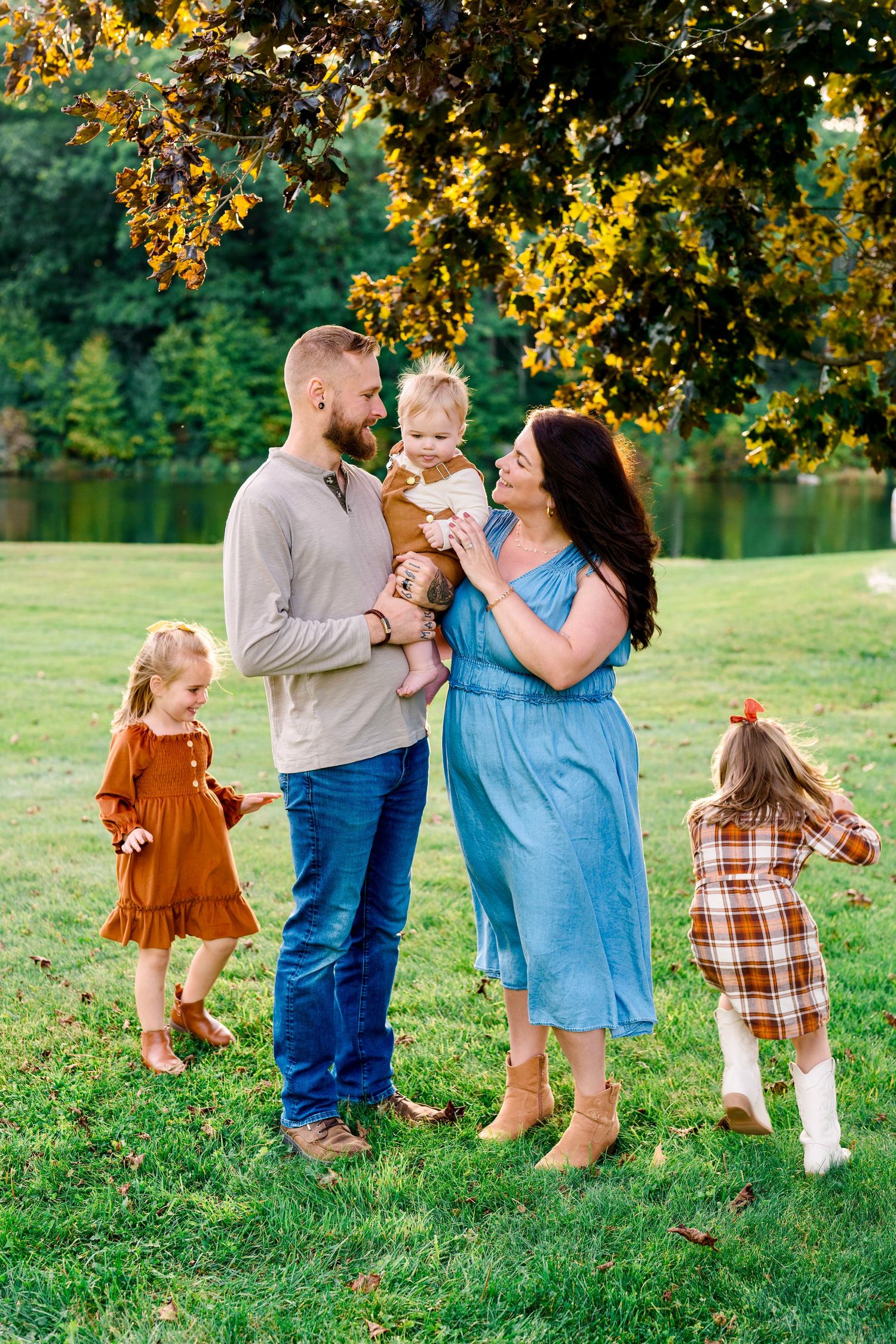 Family of five posing outdoors near a pond in autumn, parents holding toddler while two girls play nearby.