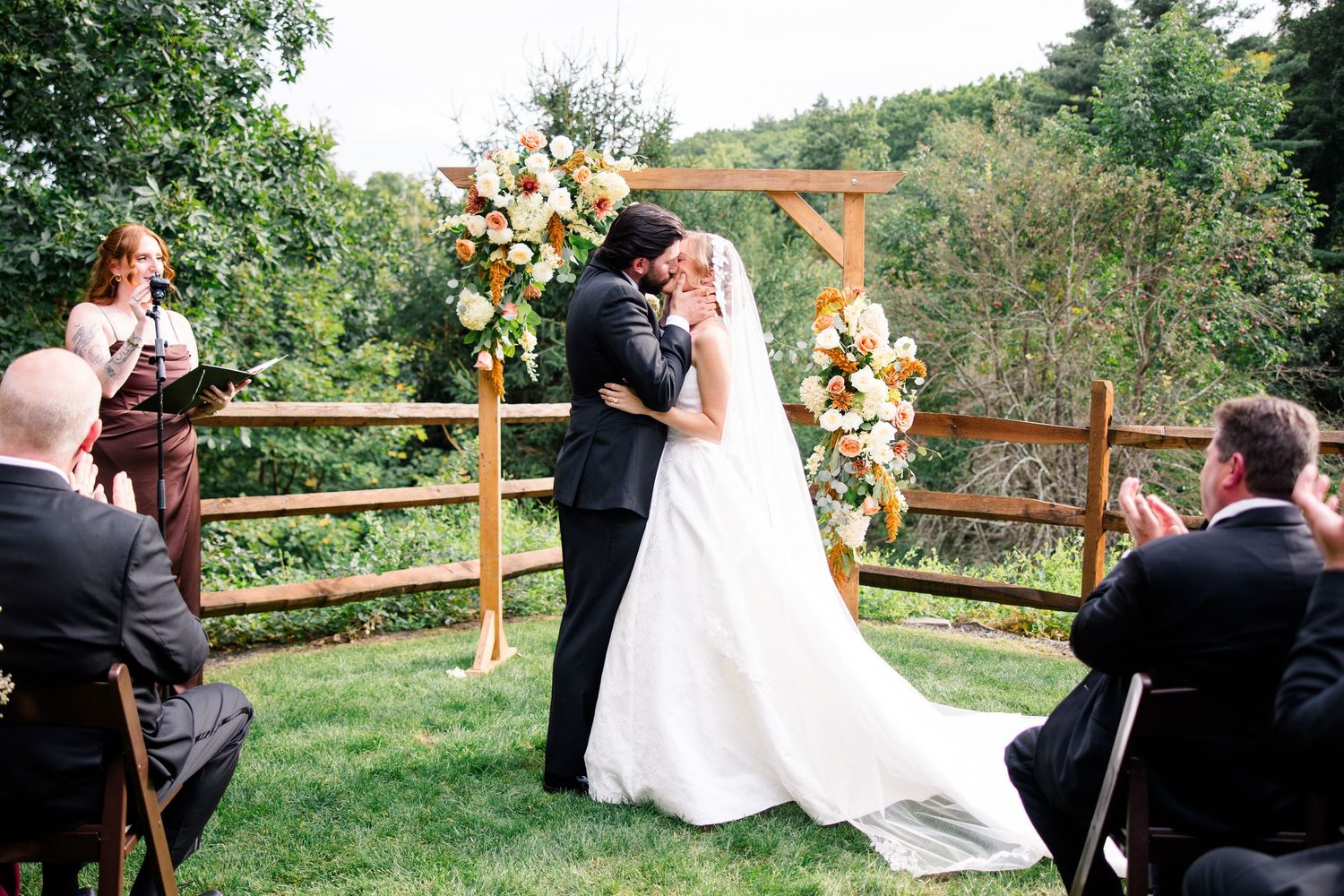 Couple shares first kiss at outdoor wedding ceremony with floral arch, wooden fence backdrop, and seated guests on green lawn.