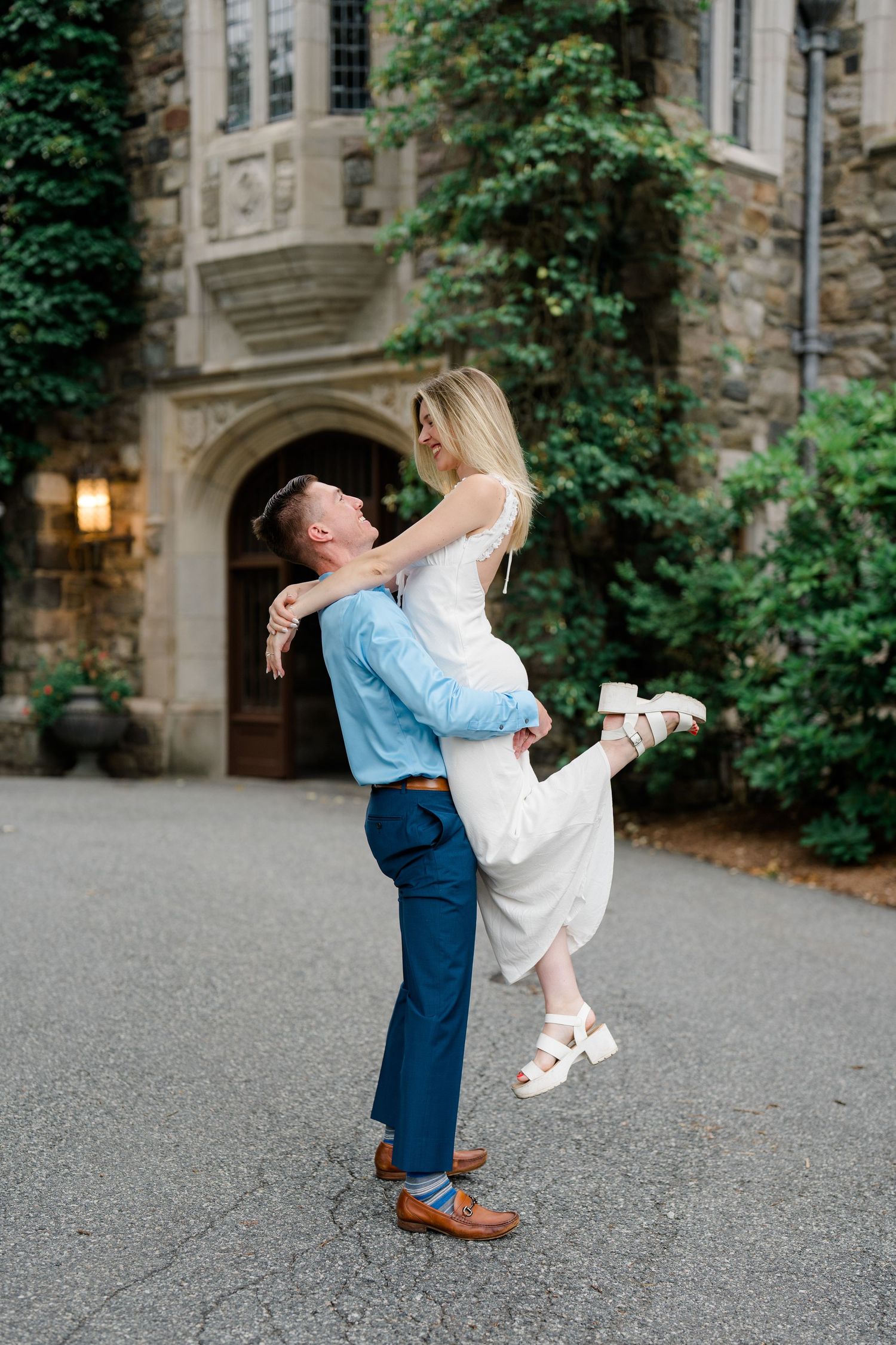 A man lifts a woman in a white dress outside a stone building covered in ivy, sharing a romantic moment.