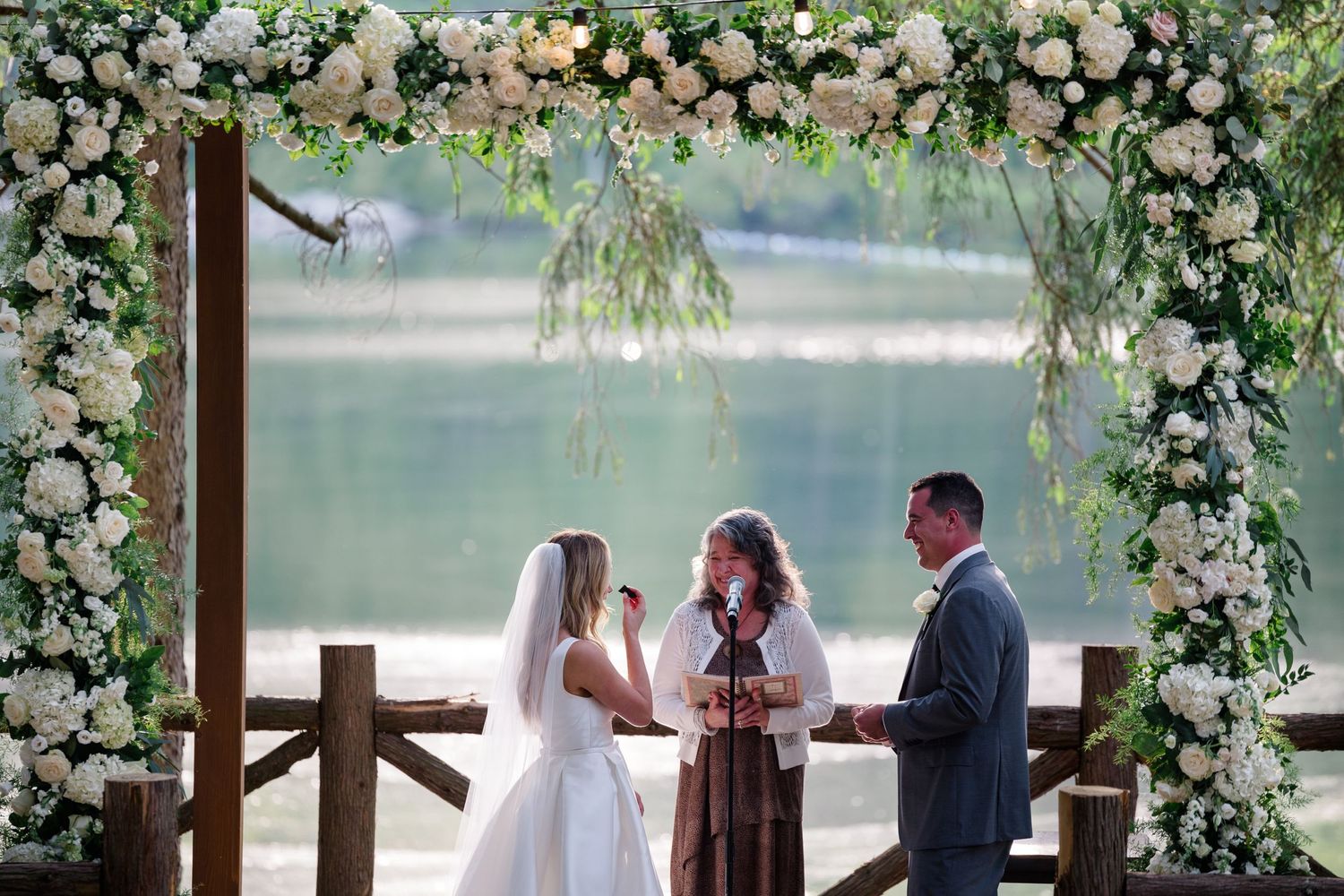 Wedding ceremony under a floral arch with white roses and greenery overlooking a serene lake.