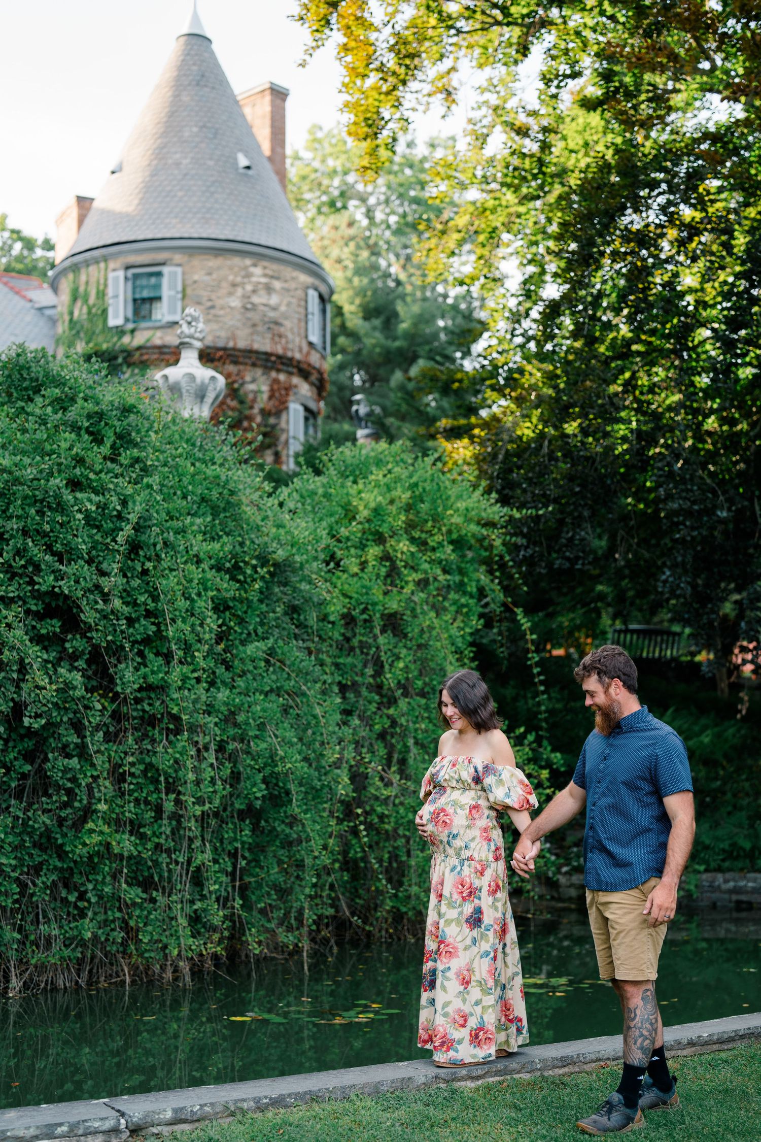 Couple holding hands by a pond with a stone turret building and lush green trees in the background.