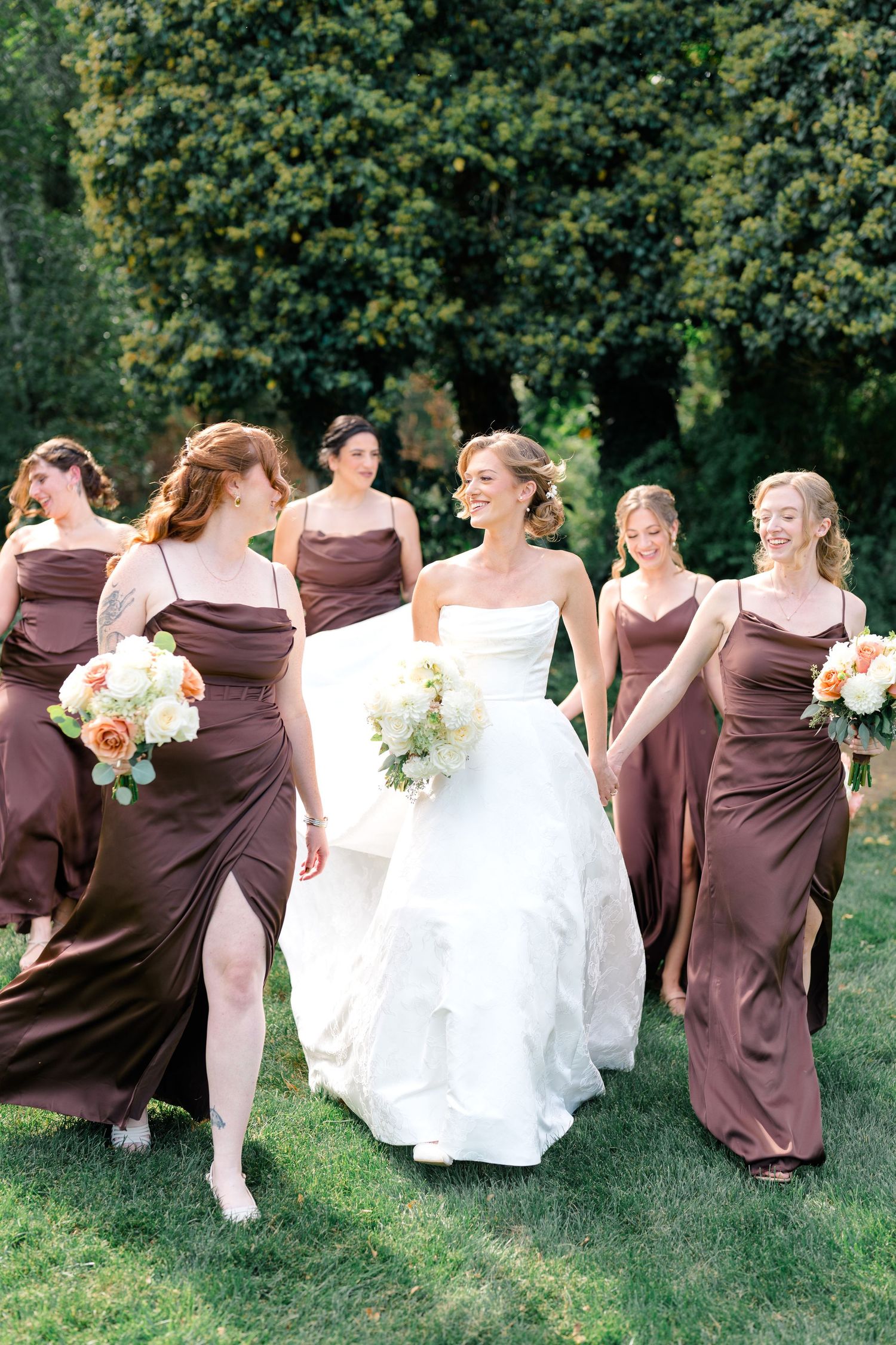 Bride in white gown walks with bridesmaids in brown satin dresses holding floral bouquets on green lawn.