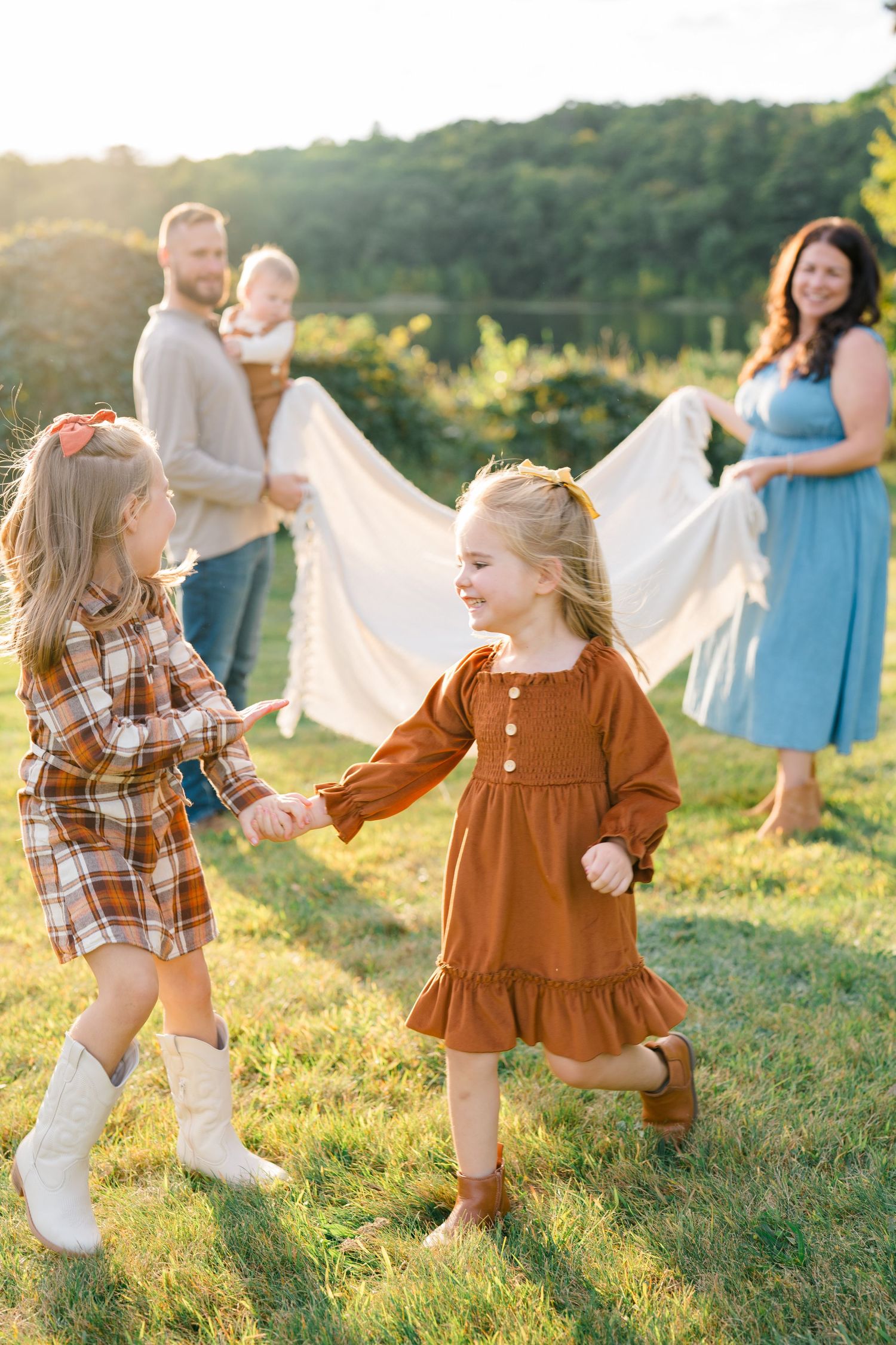 Two young girls in fall dresses hold hands outdoors while parents hold a white blanket in golden sunlight.