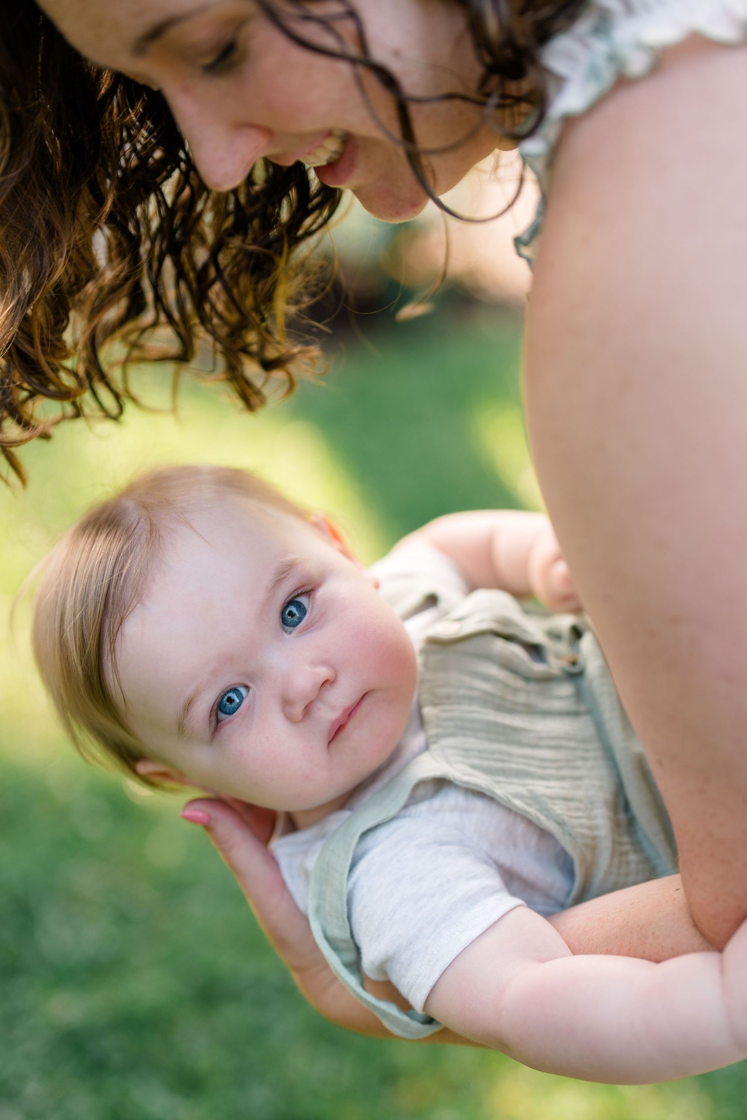 A smiling mother with curly hair holds her blue-eyed baby outdoors on a sunny day with green grass background.