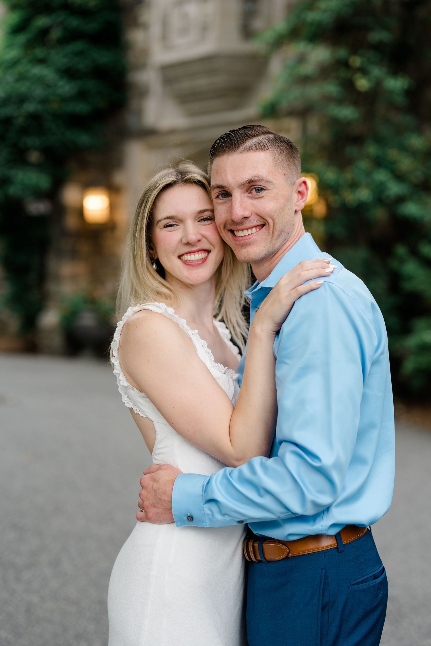 Smiling couple embracing outdoors near a stone building, woman in white lace dress and man in blue shirt.