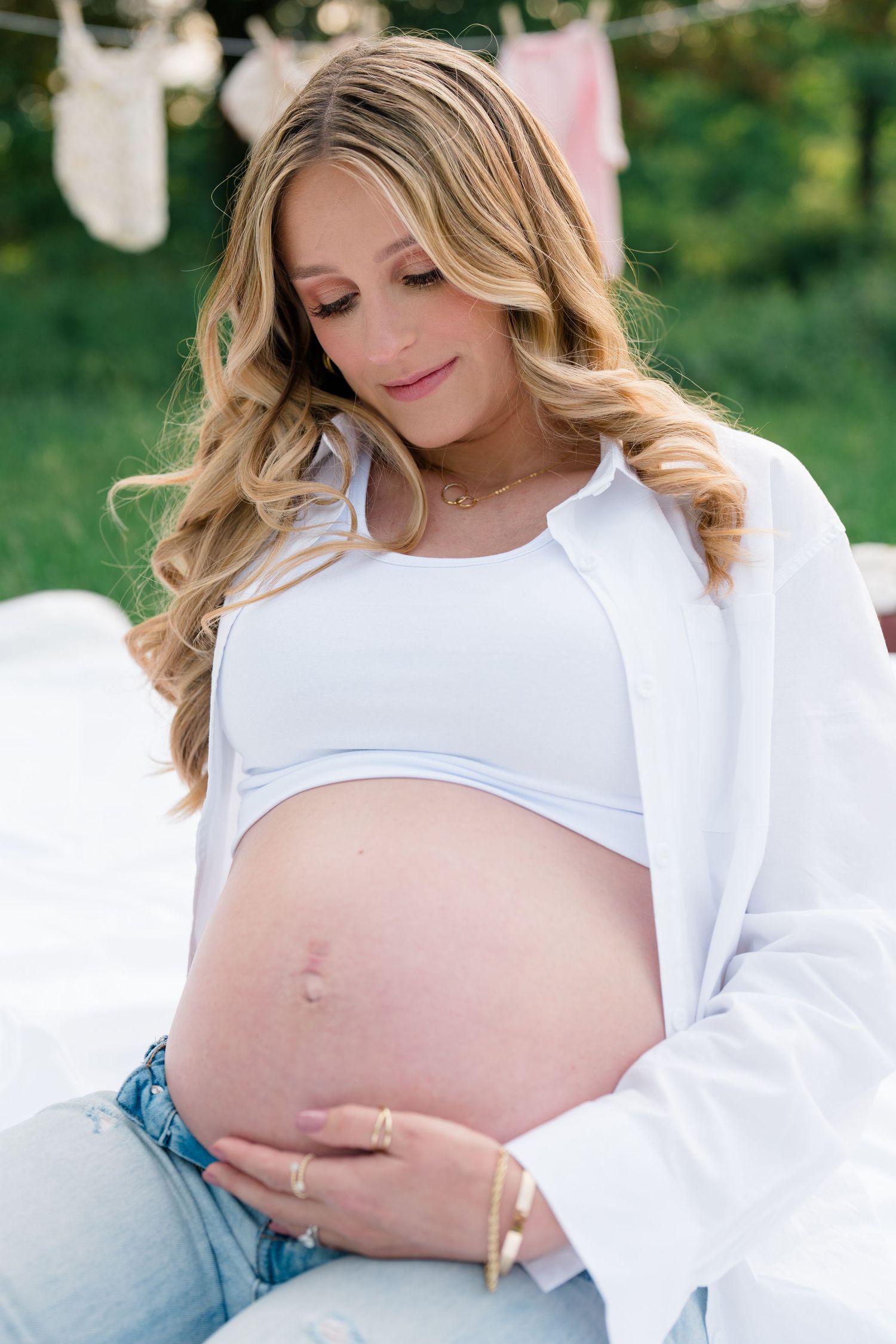 Expectant mother in white top and jeans caresses her baby bump while smiling in an outdoor maternity photo session.