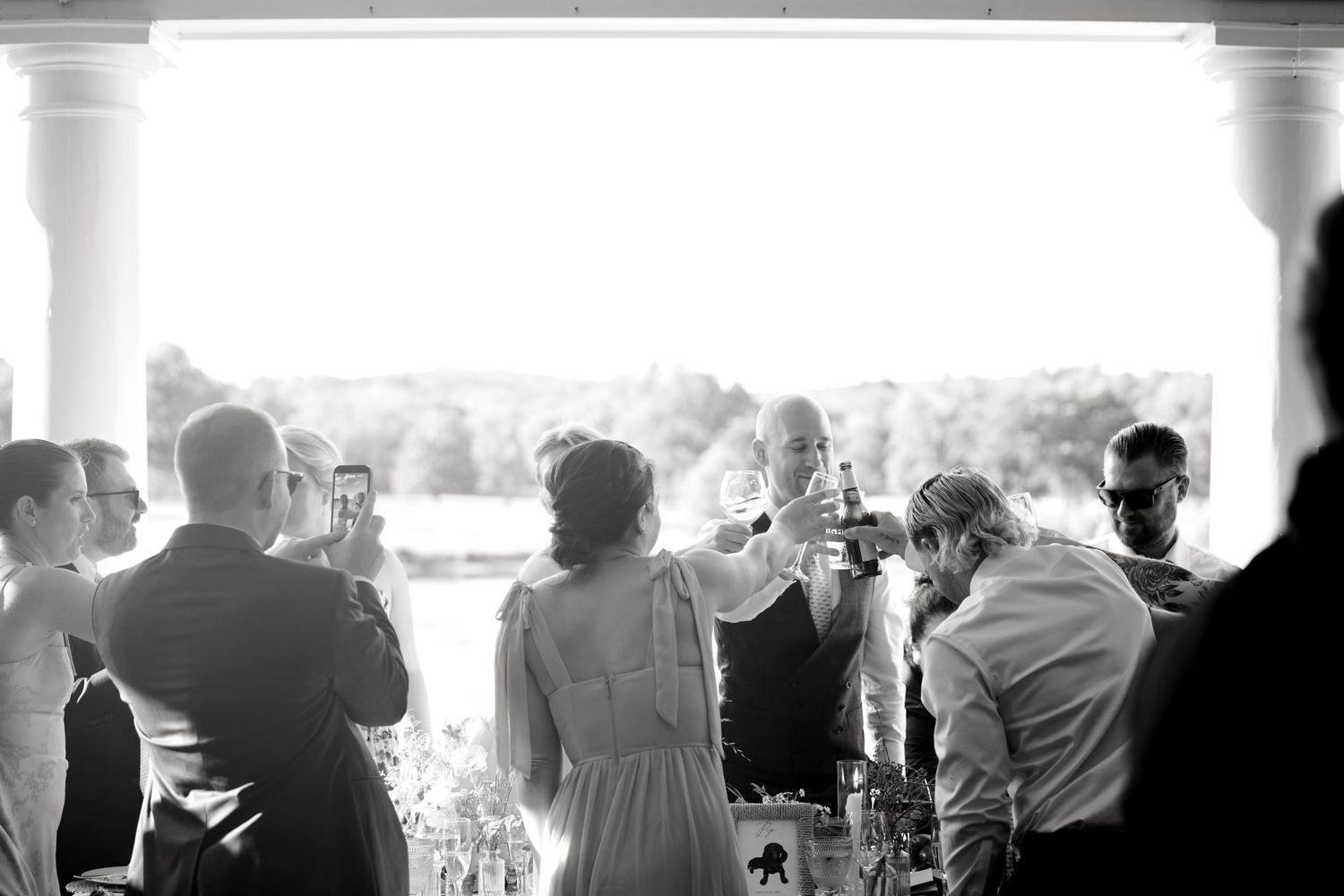 Black and white photo of wedding guests raising champagne glasses in a toast on a sunlit porch.