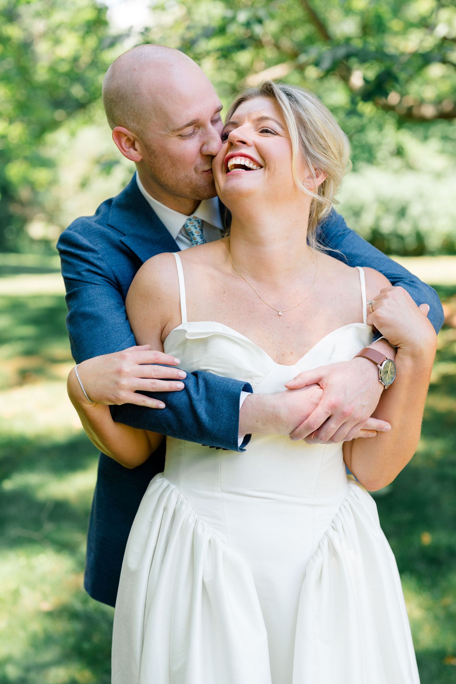 Joyful bride in white dress laughs as bald groom in navy suit embraces her from behind in garden setting.