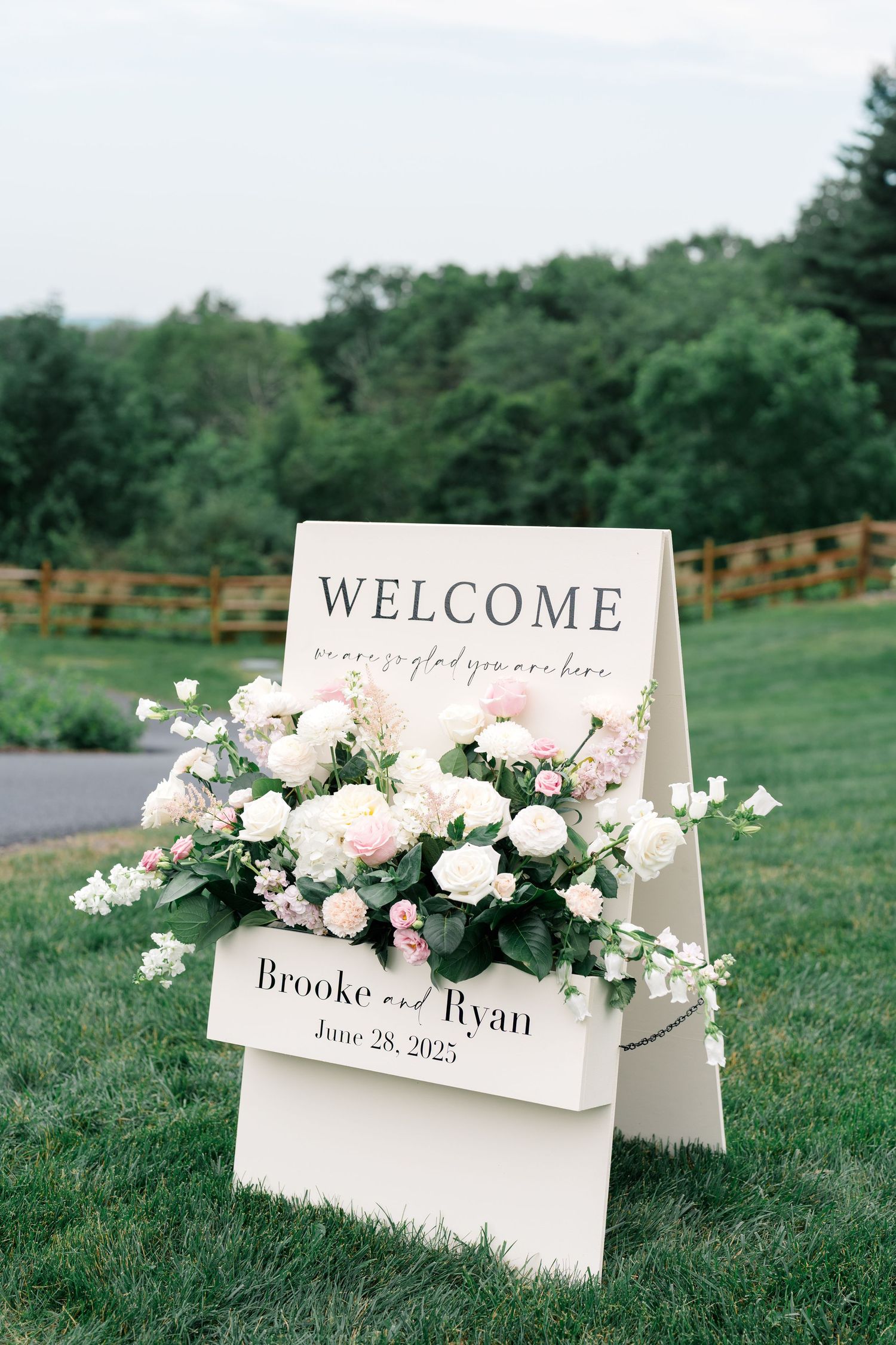 White welcome signs with floral arrangements displayed on grass at outdoor wedding.