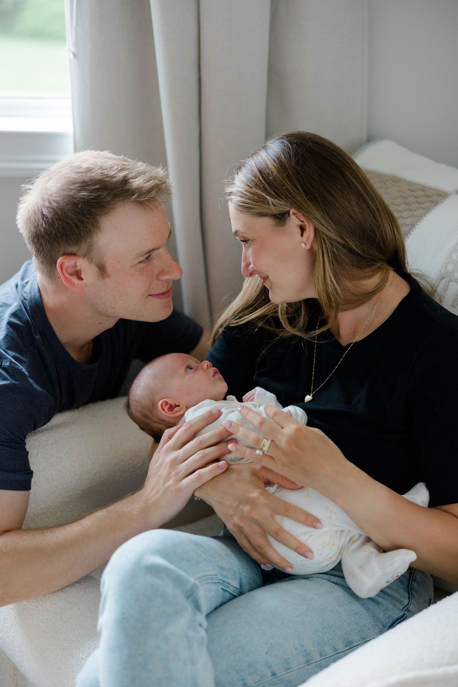 Parents cuddling with their newborn on a comfortable gray couch in natural light.