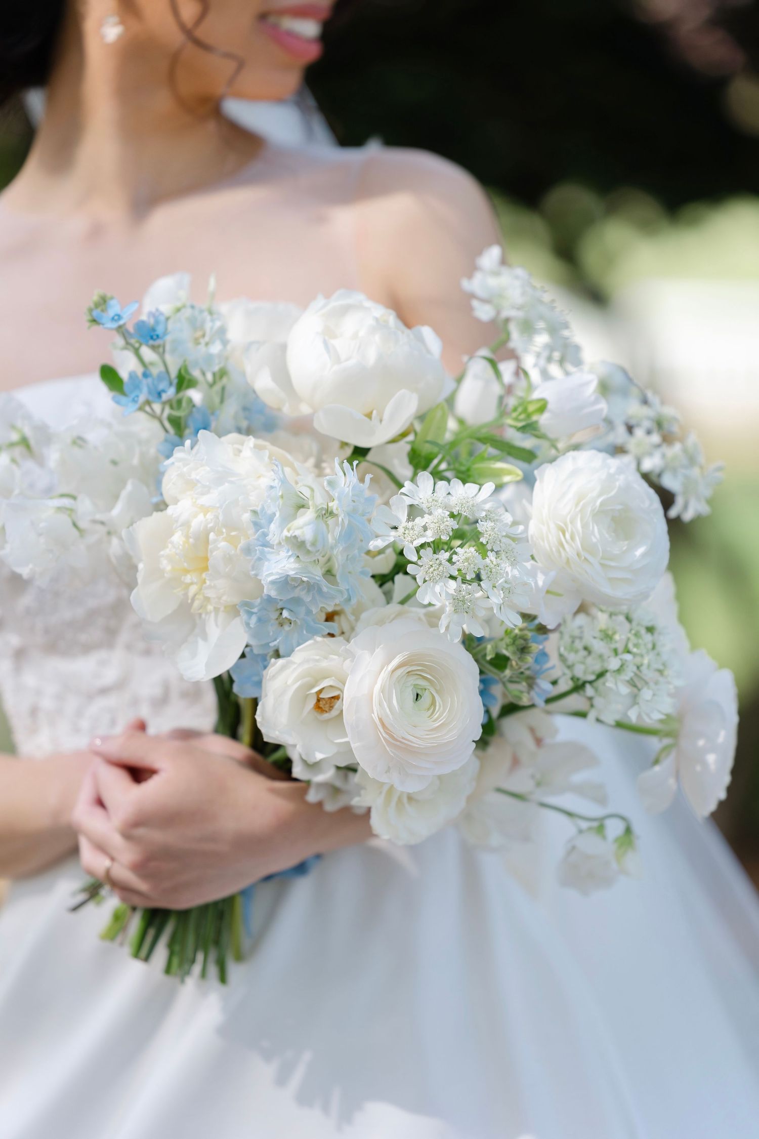 Bride holding a lush wedding bouquet of white peonies, ranunculus, and soft blue hydrangeas in an elegant bridal gown.