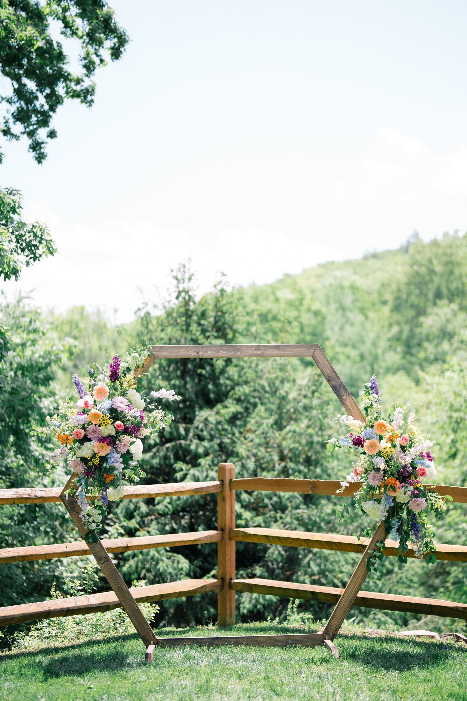 Rustic wooden hexagon wedding arch decorated with colorful floral arrangements overlooking a scenic hillside garden.