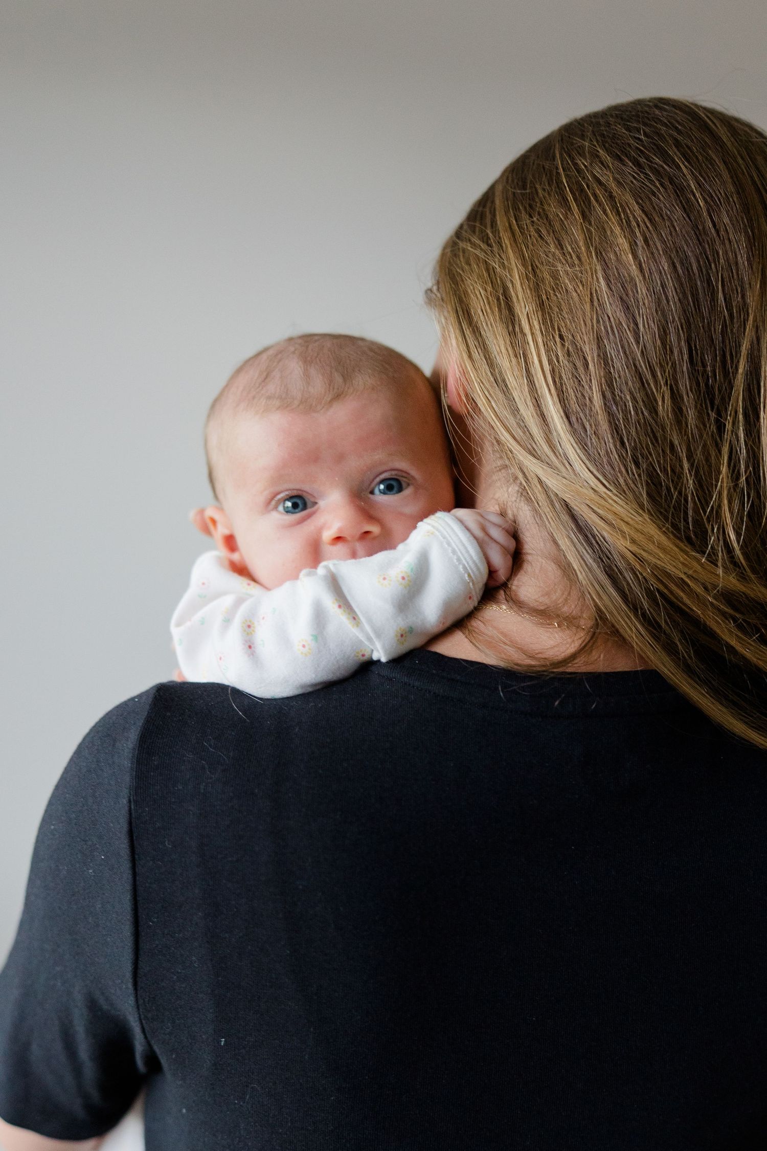 Baby in white onesie rests on shoulder against black shirt with blonde hair visible.