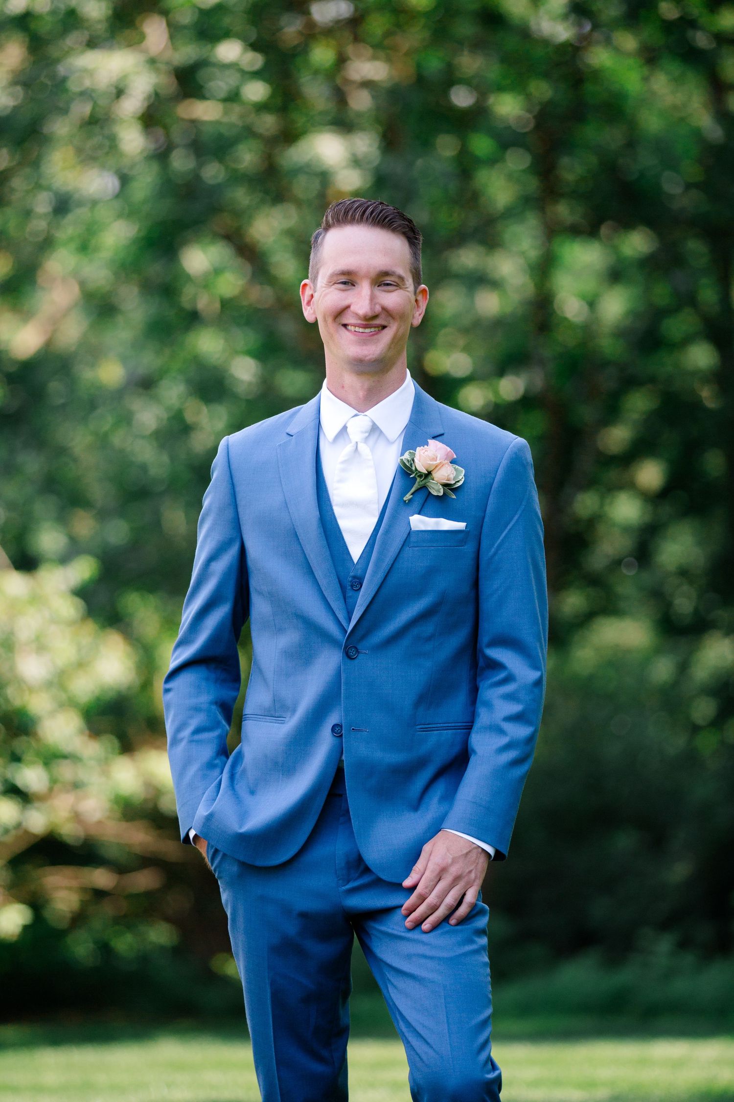 Groom in light blue suit with boutonniere smiles for a portrait in garden setting.