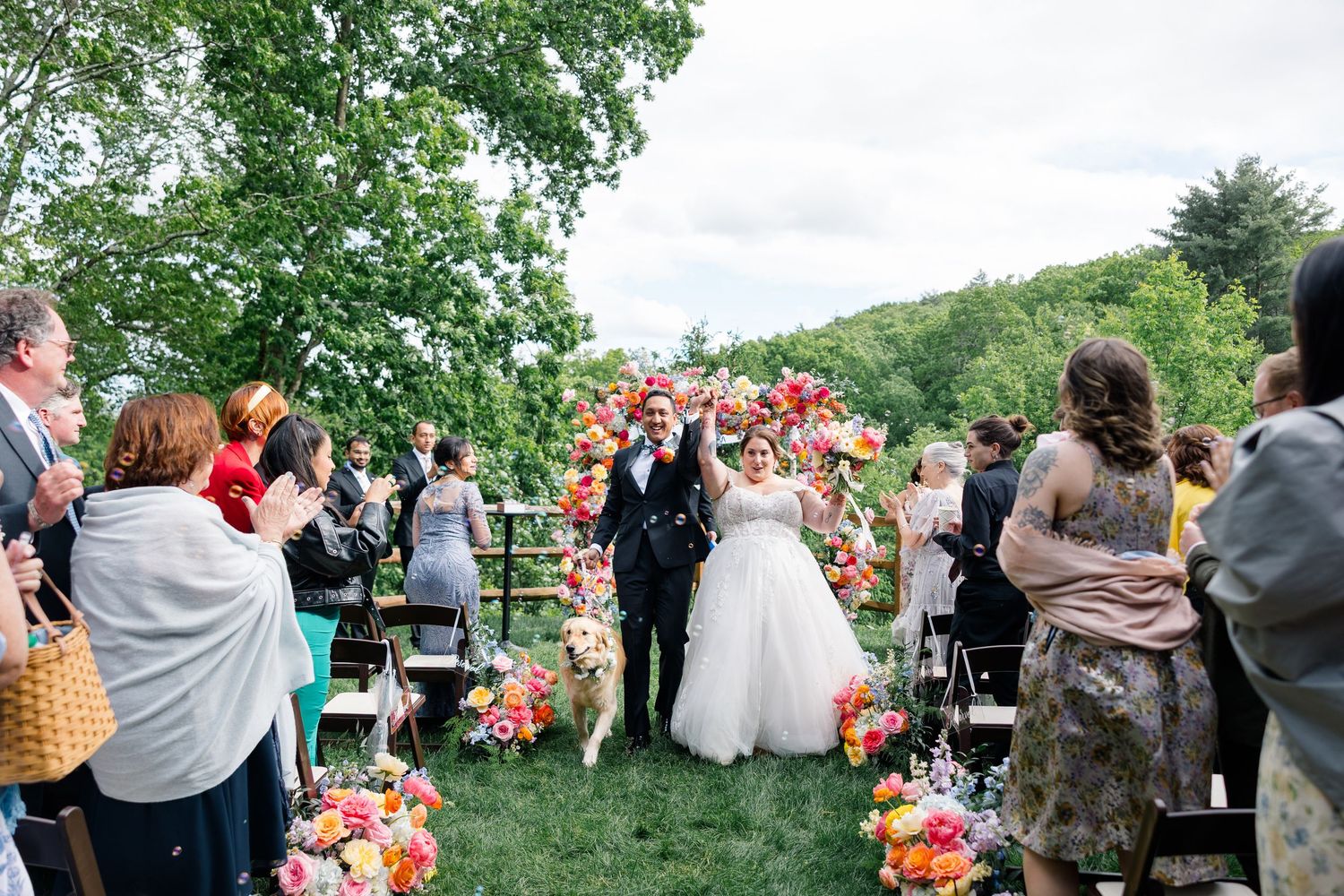 Outdoor wedding ceremony with guests watching couple exchange vows surrounded by vibrant coral flower arrangements.