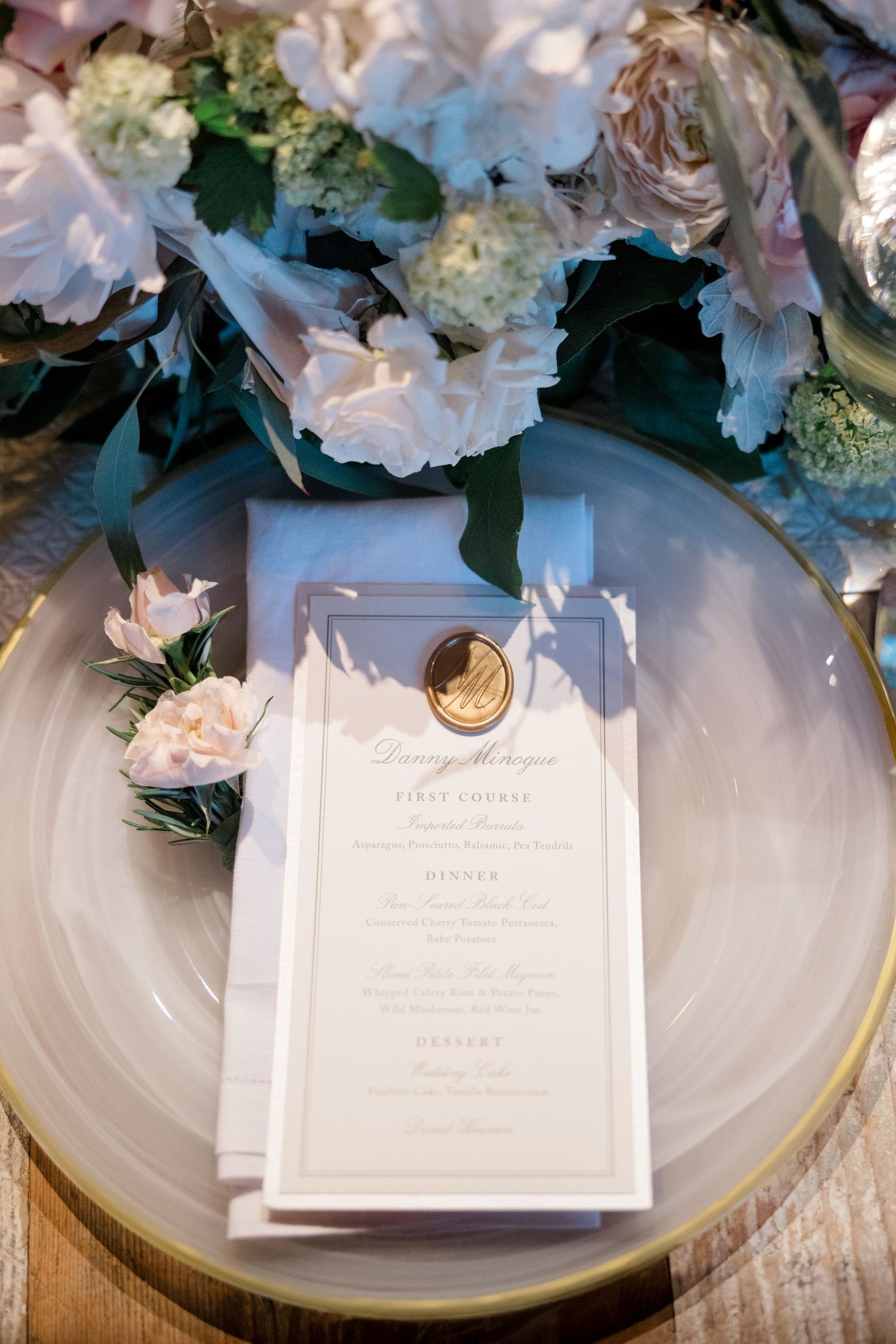 An elegant wedding menu card with gold wax seal sits on a plate surrounded by white and pink floral arrangements.