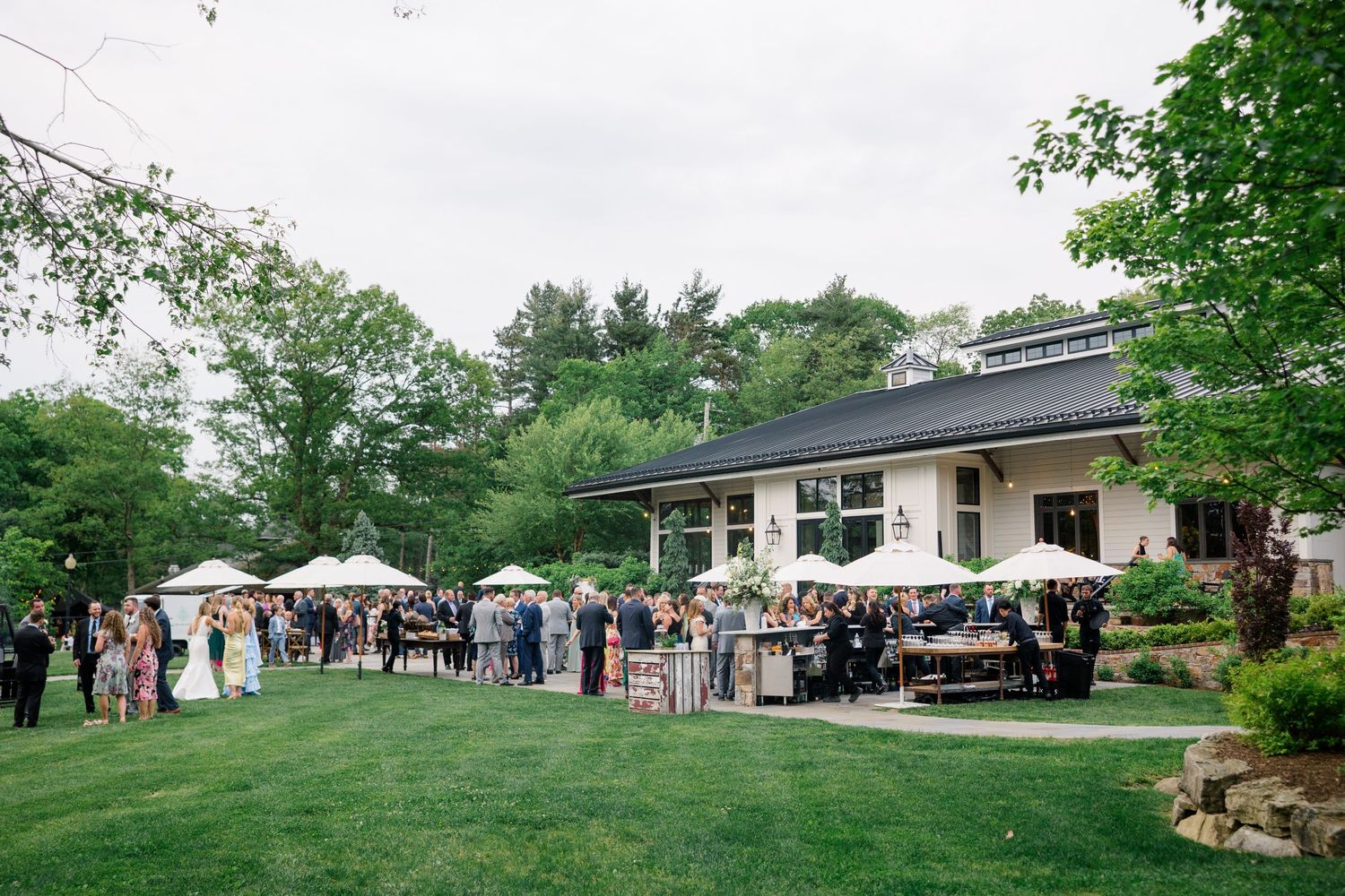 Wedding guests mingle on a lush lawn outside a venue with tents and tables set up for an outdoor reception.