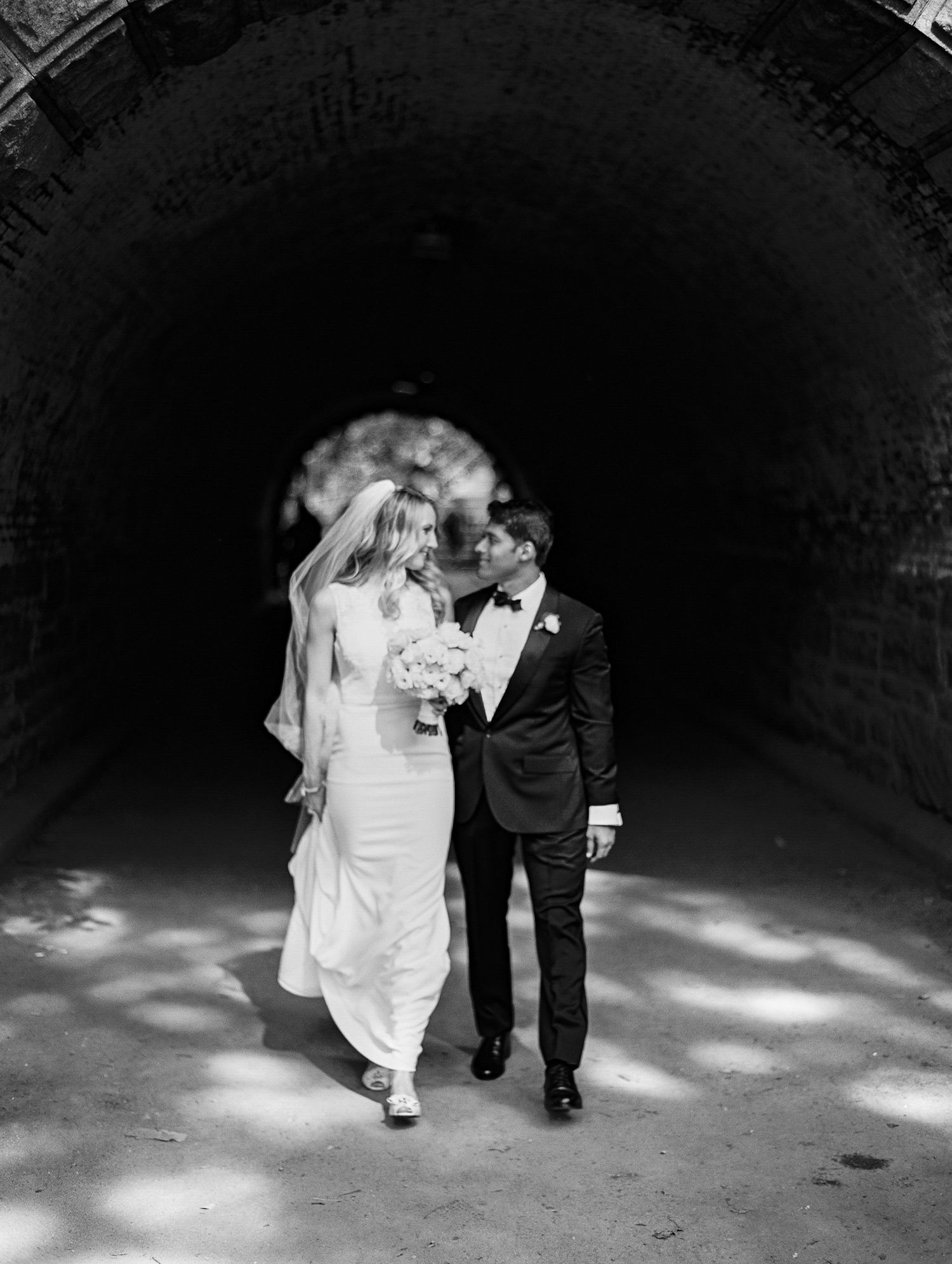 Black and white wedding photo of couple walking through a dark tunnel with dramatic lighting.
