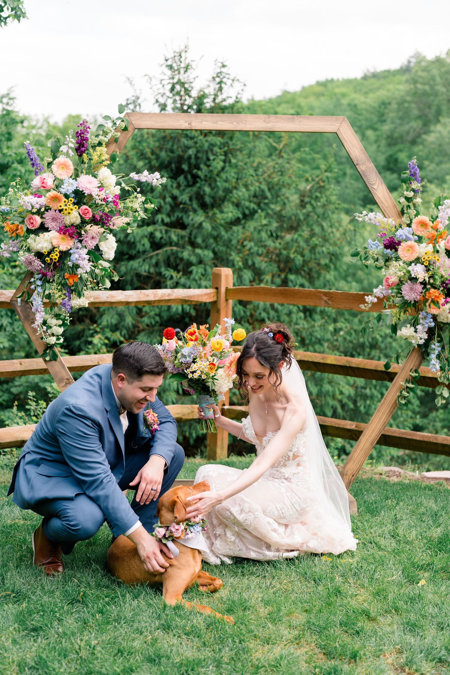A couple interacts with their dog under a floral arch during their wedding ceremony.