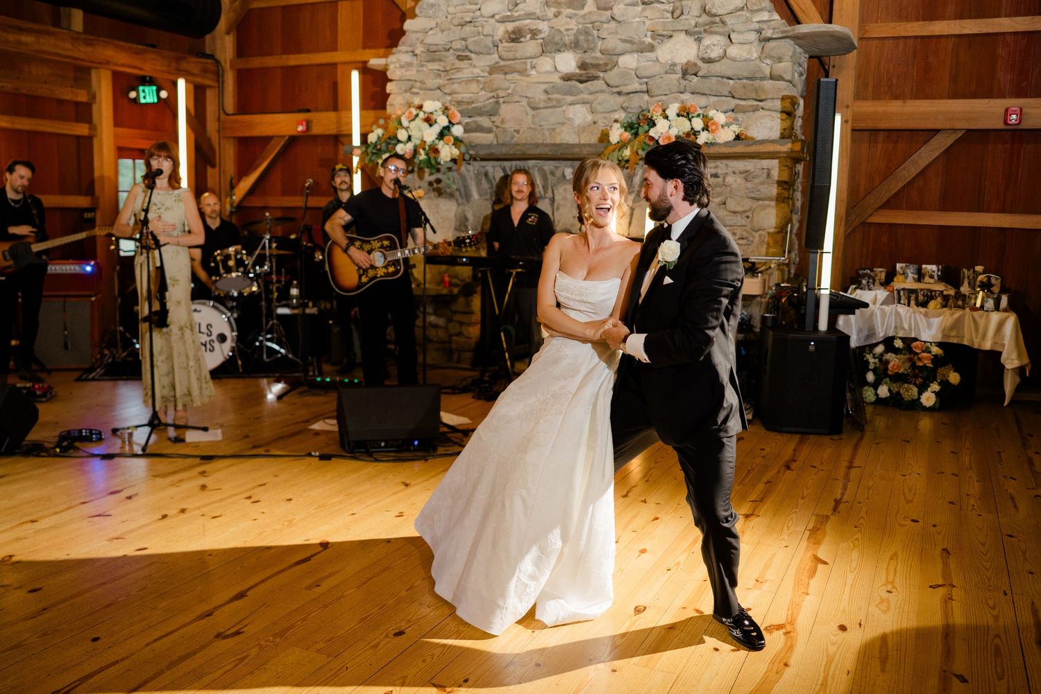 Bride and groom share first dance in rustic barn venue with live band performing and stone fireplace backdrop.