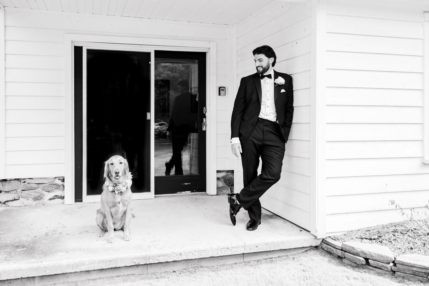 Groom in tuxedo leaning against white house exterior beside a golden retriever dog in black and white wedding photo.