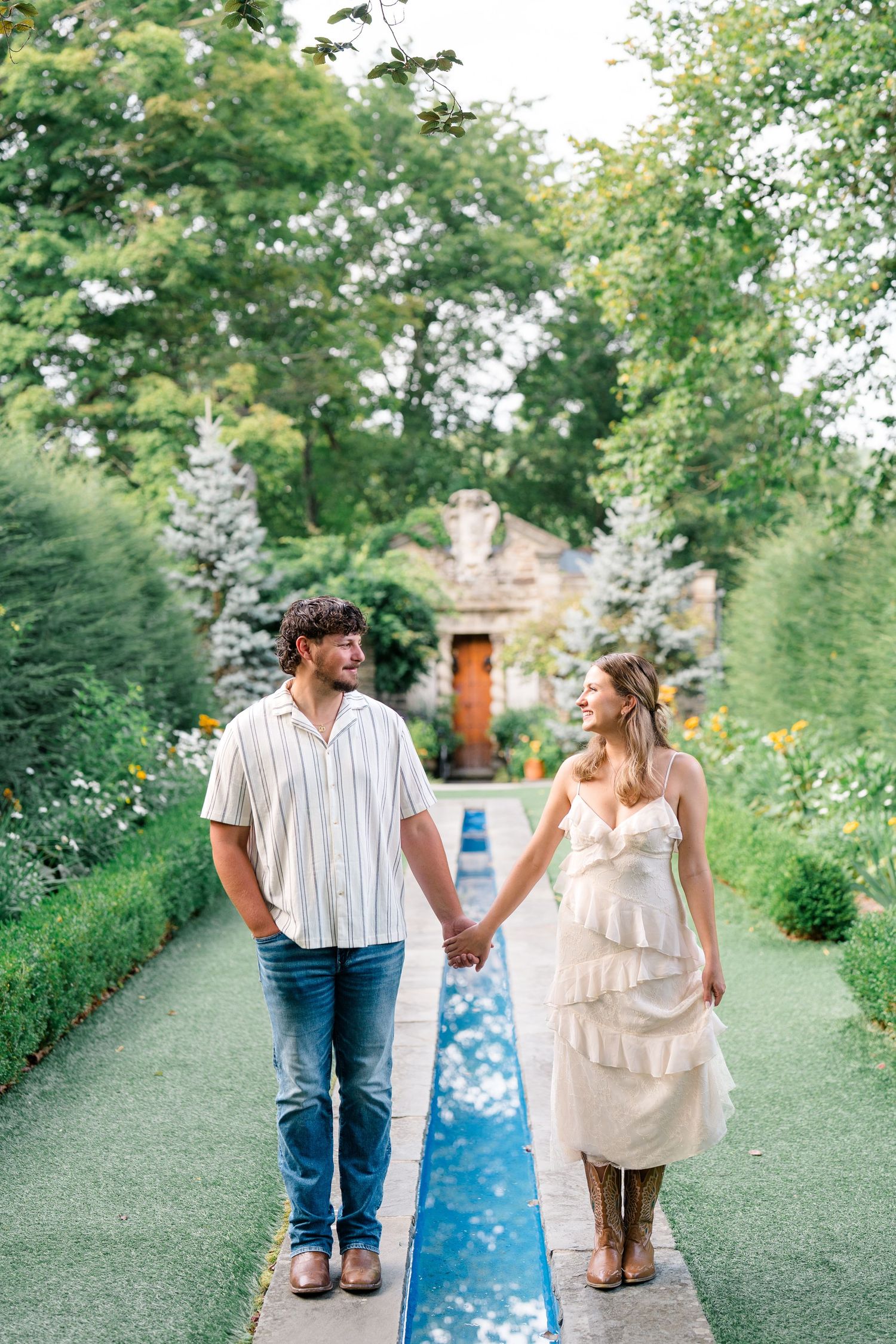 Couple holding hands walking along a garden path with lush greenery and a charming cottage in the background.