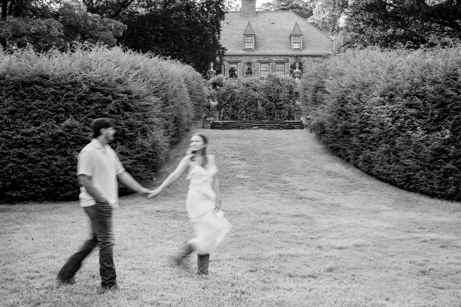 Black and white photo of a couple holding hands and walking through manicured hedges with a historic manor house behind them.
