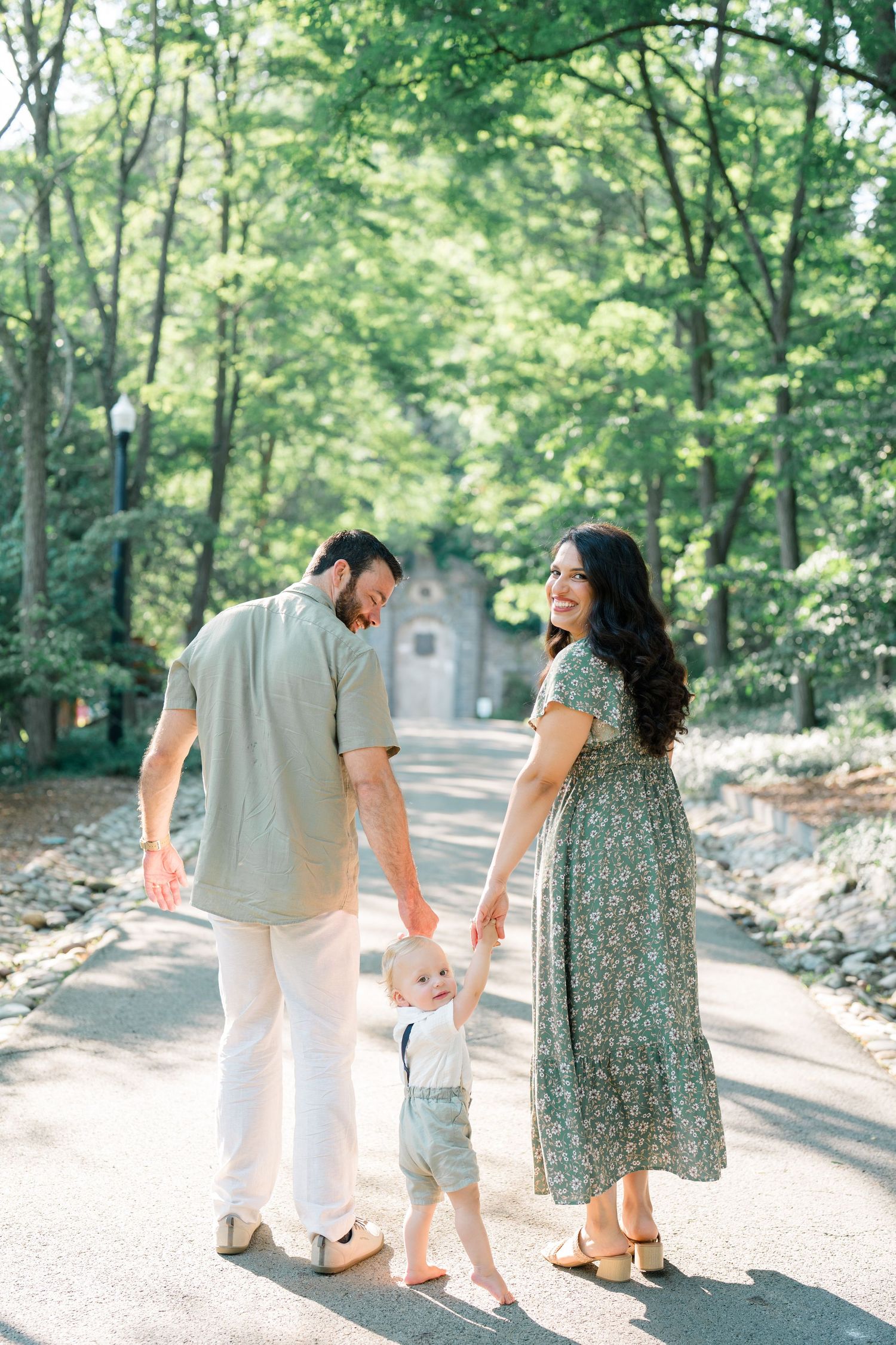 A young family of three walks hand-in-hand on a tree-lined path, mom in a floral dress, dad in casual attire.