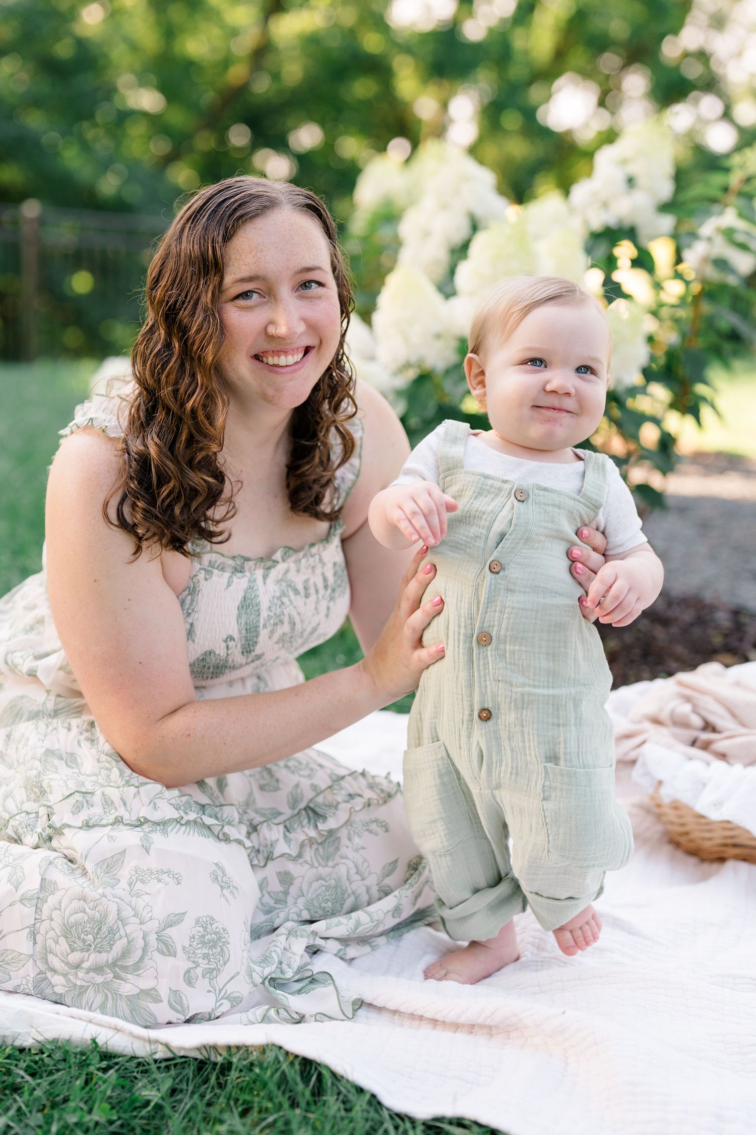 A smiling mother in a floral dress sits on a blanket outdoors with her happy baby in a green romper.