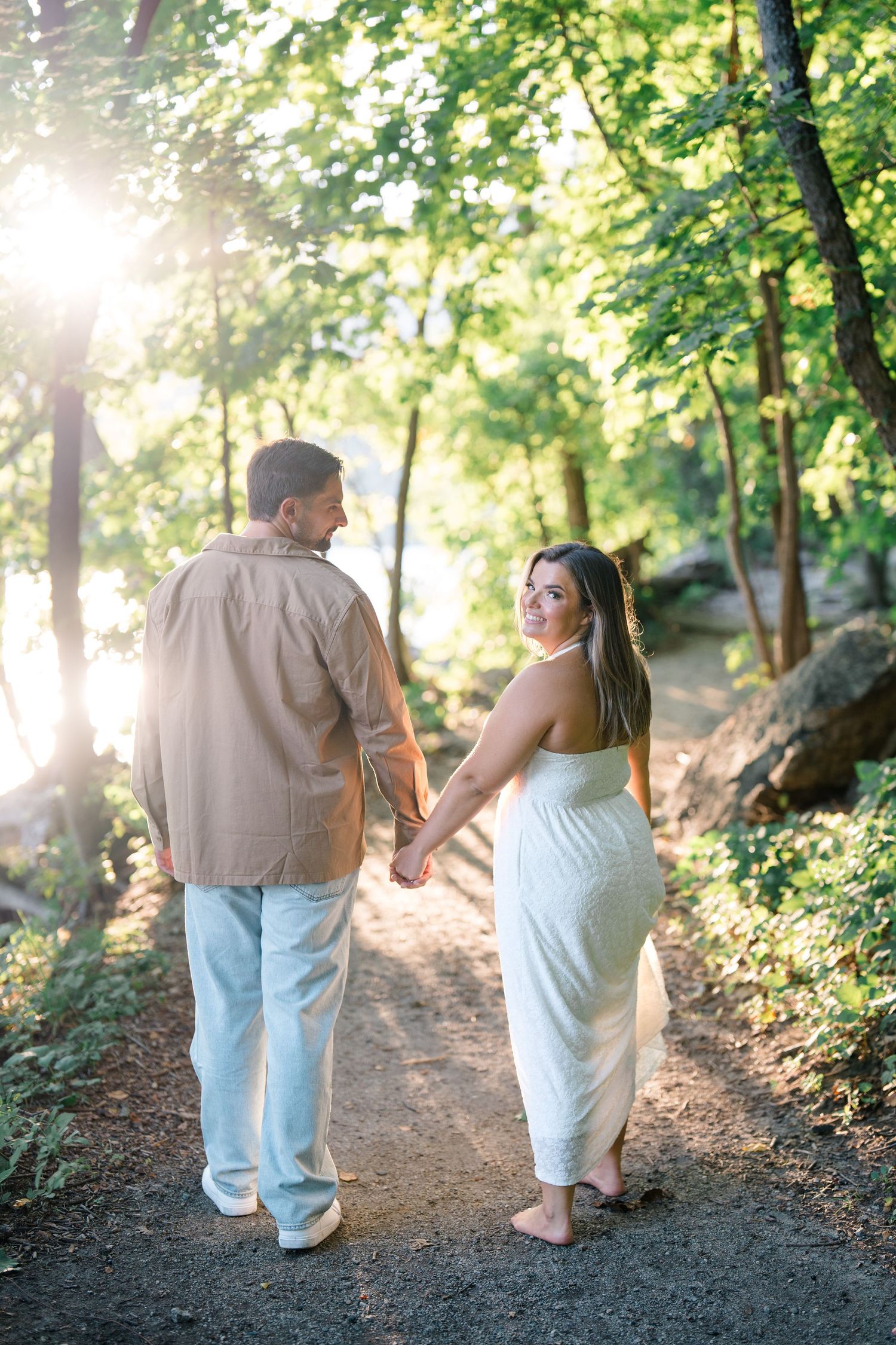Couple holding hands walking on a sunlit forest path, woman in white dress looks back, golden hour light filters through trees.