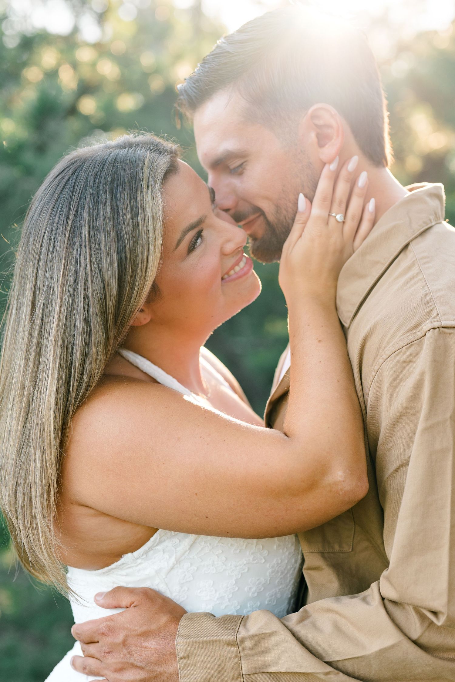 Couple sharing a romantic embrace outdoors, woman smiling with engagement ring visible, bathed in golden sunlight.