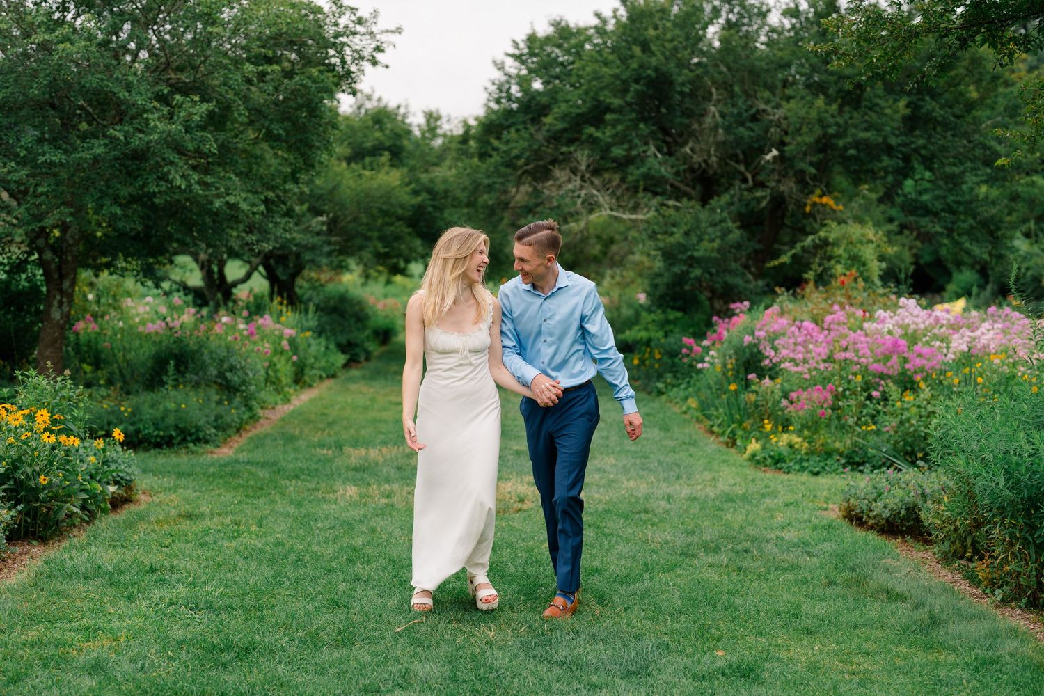 Couple walking hand-in-hand through a lush garden path surrounded by colorful flowers and green trees.