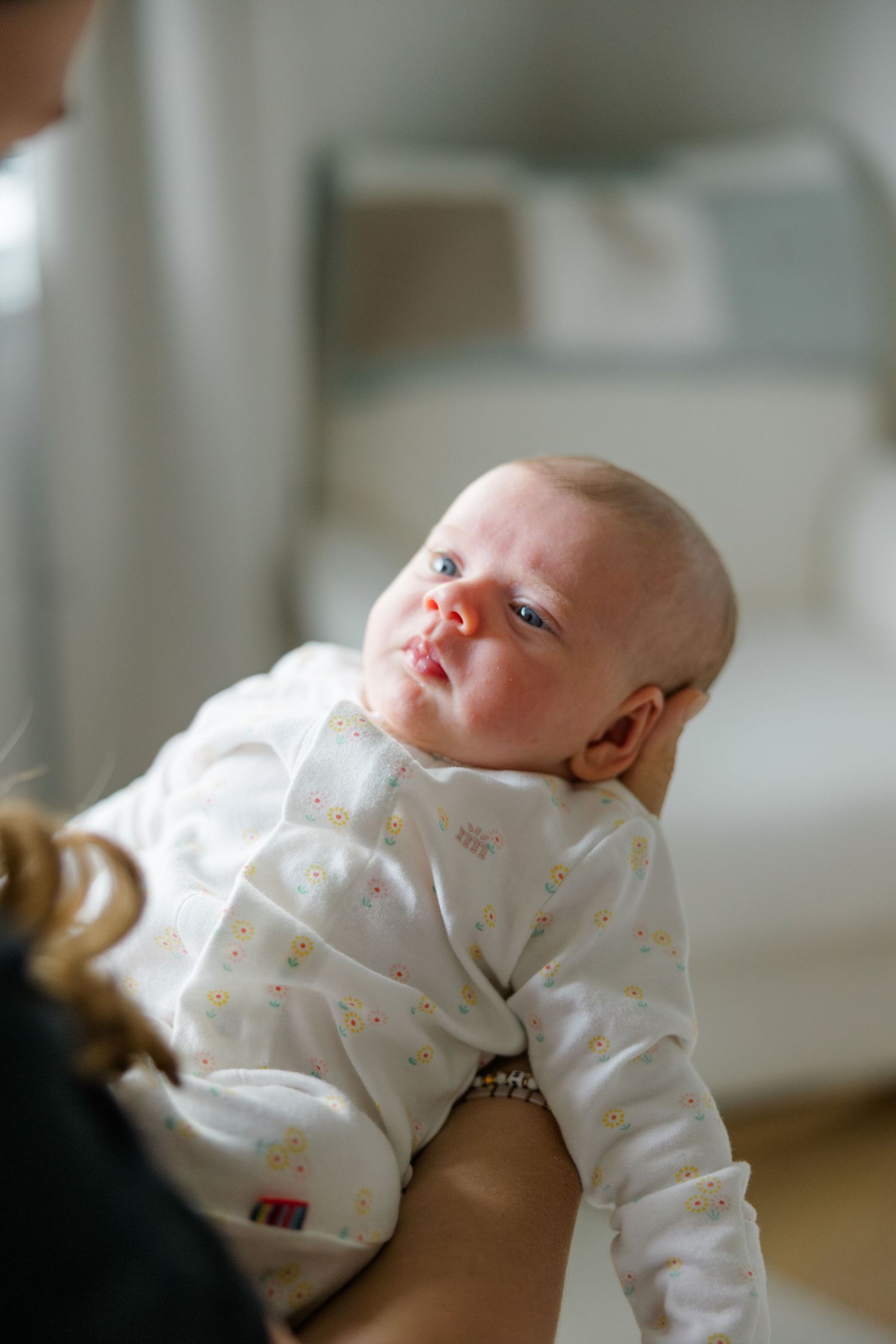 Baby wearing white patterned onesie looks up while being held.