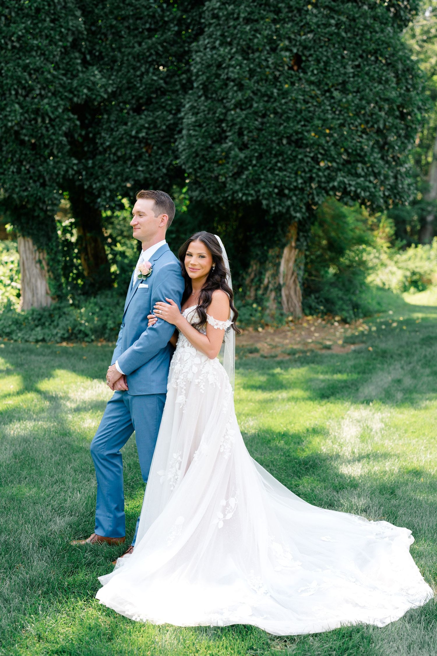 Classic wedding portrait of couple in blue suit and white gown posing back-to-back outdoors.