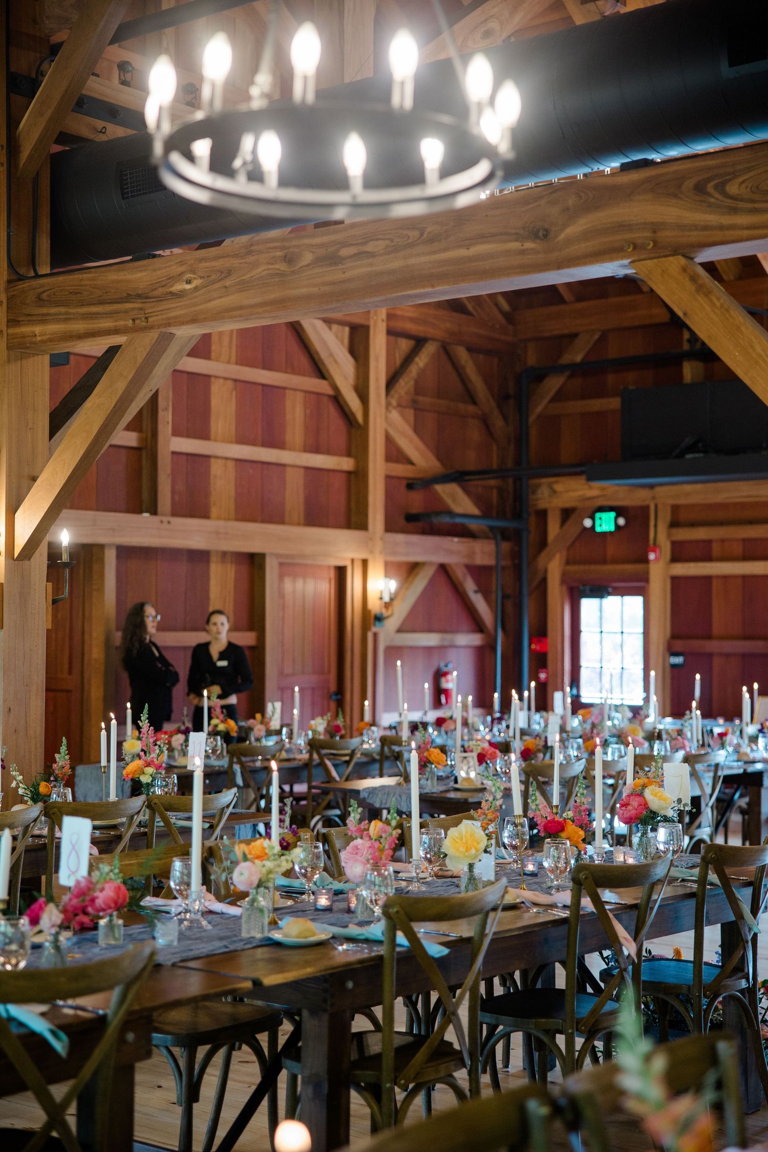Elegant barn reception venue with rustic chandelier, wooden beams, and tables decorated with floral centerpieces and candles.