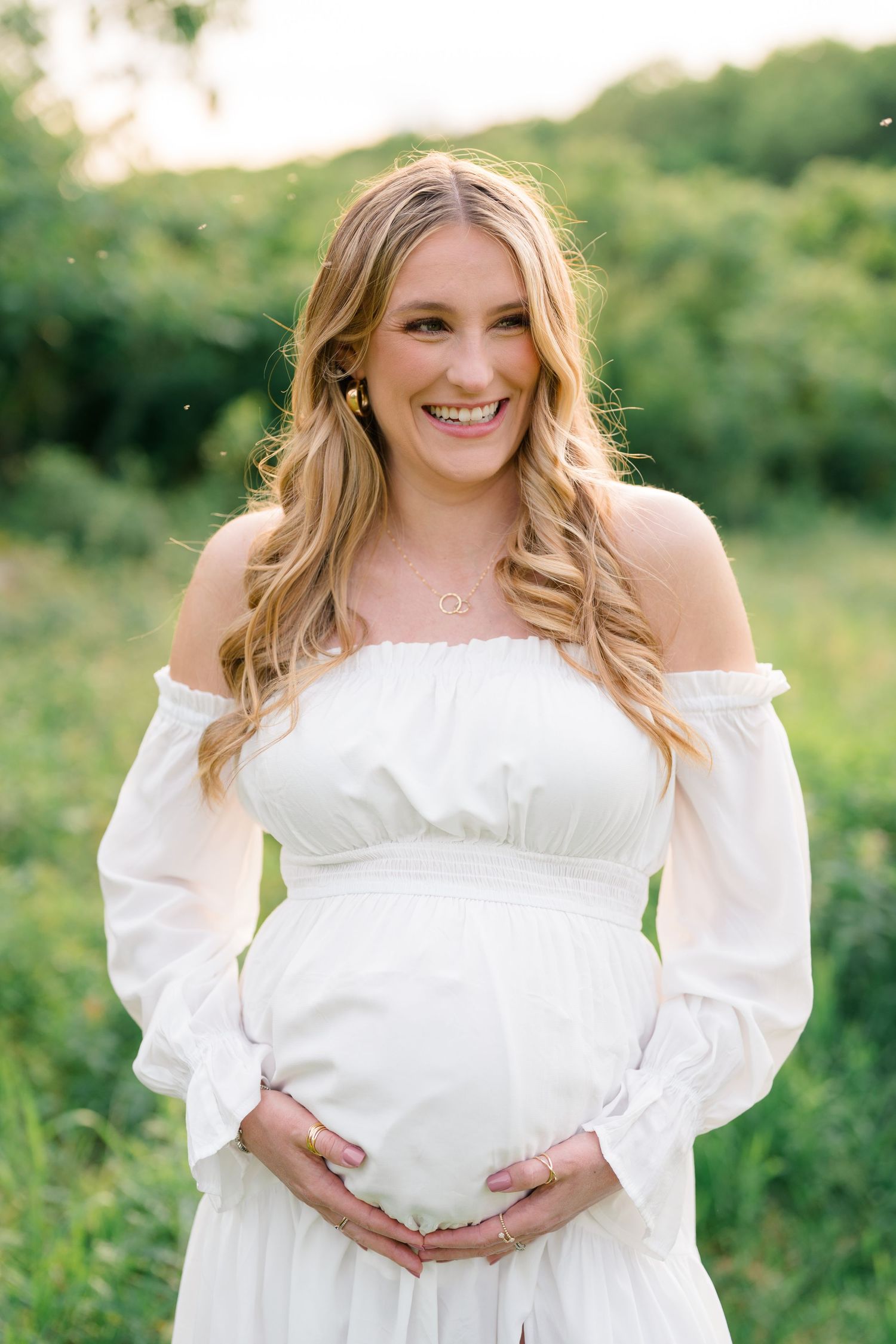 Maternity portraits in a white off-shoulder dress against lush green outdoor backdrop.