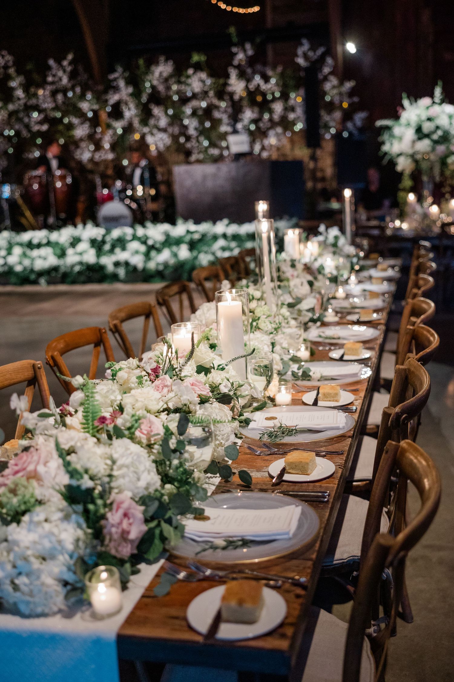 Elegant wedding reception table with floral centerpieces, candles, and wooden chairs in a rustic venue.