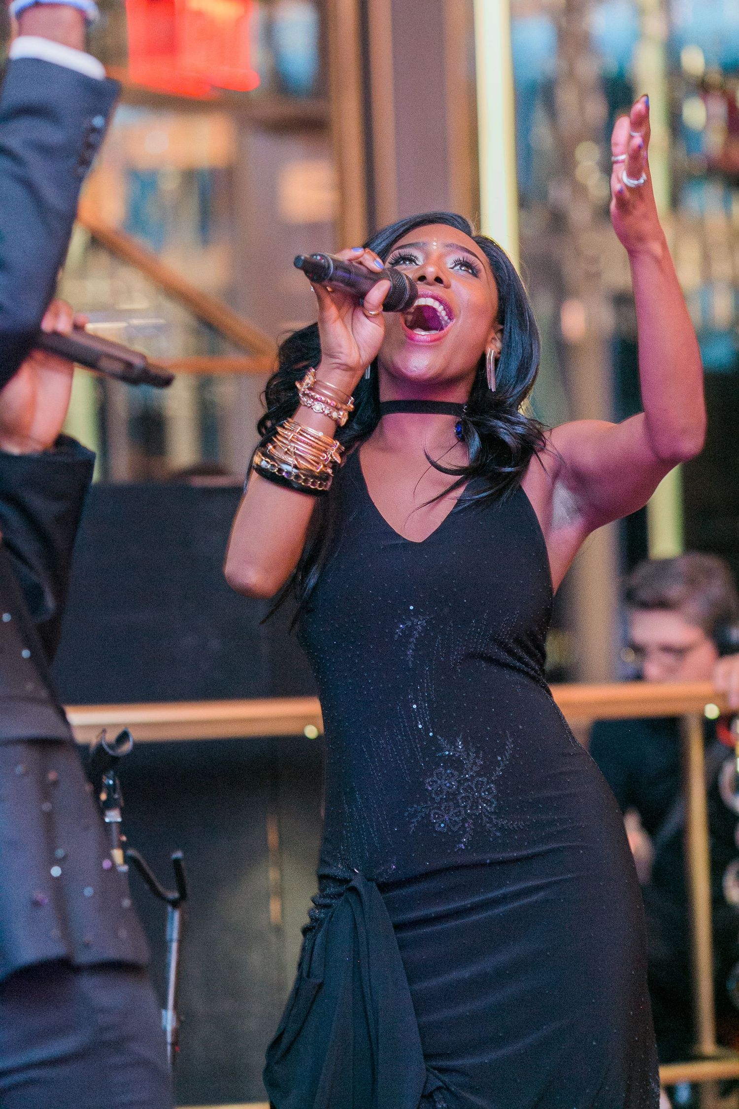 Singer performing passionately on stage wearing a black dress and bracelets with dramatic lighting in the background.