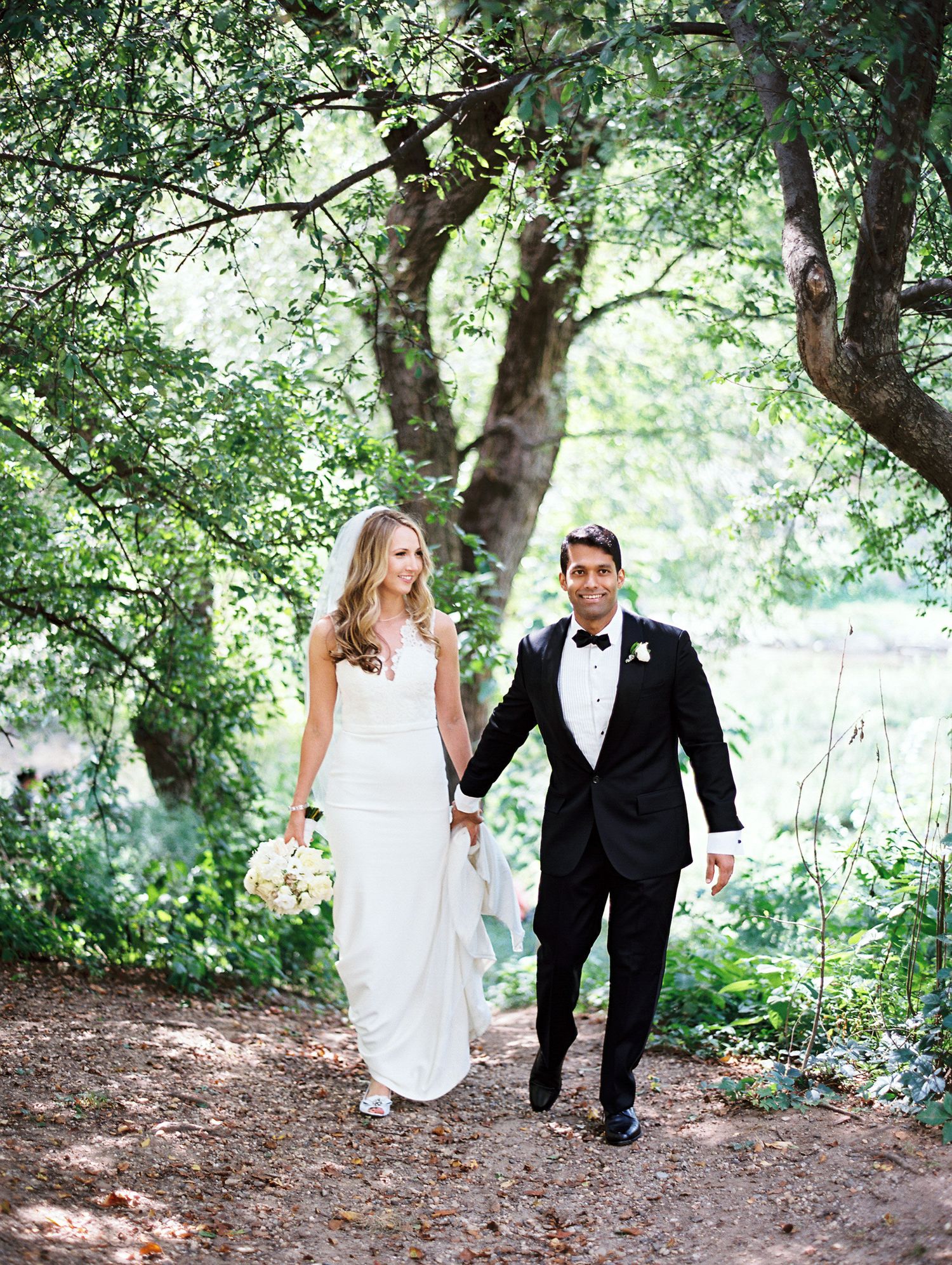 A couple in wedding attire walk hand in hand along a shaded forest path beneath tall trees and dappled sunlight.