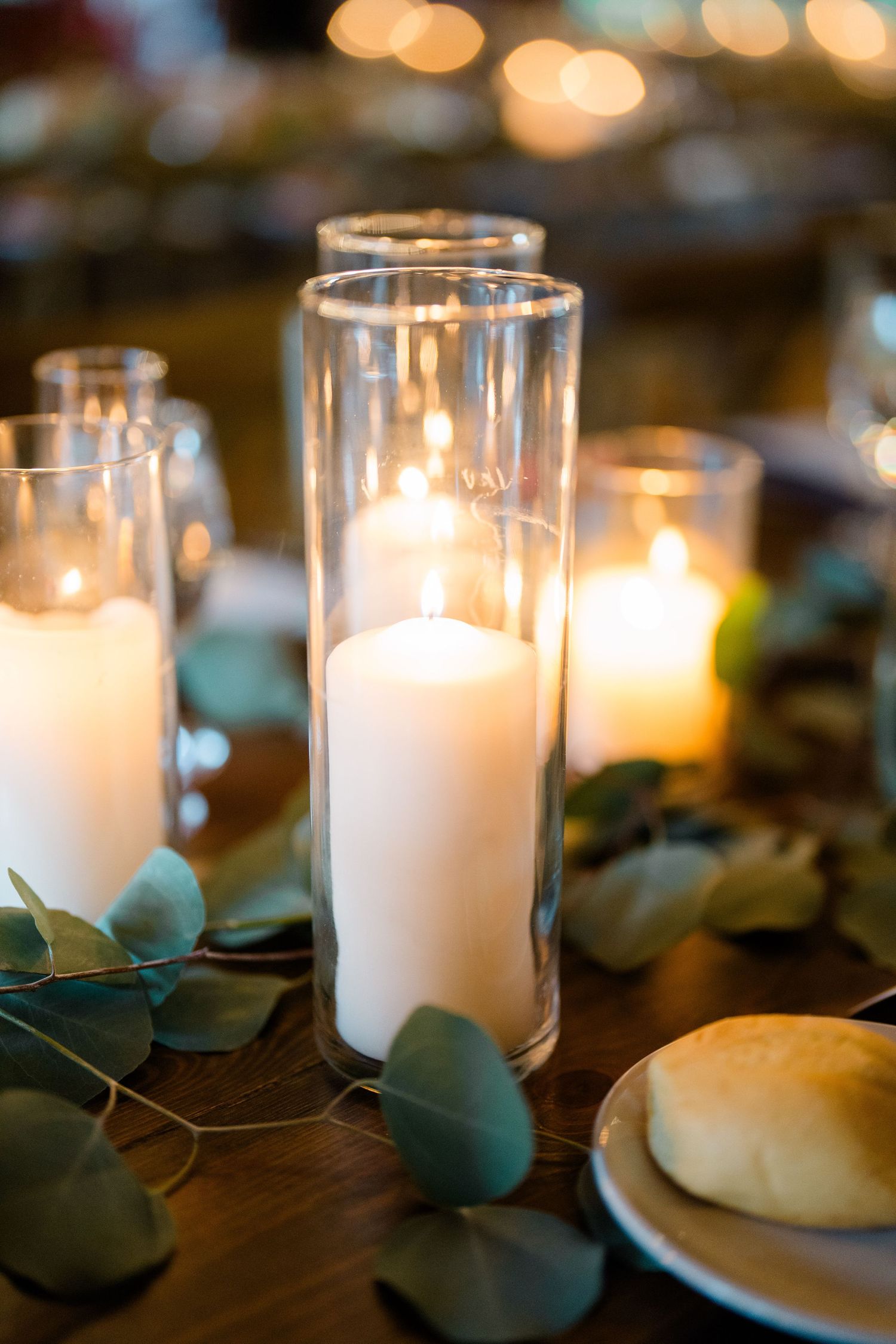 Elegant wedding reception centerpiece with white pillar candles in glass cylinders surrounded by eucalyptus greenery on wooden table.